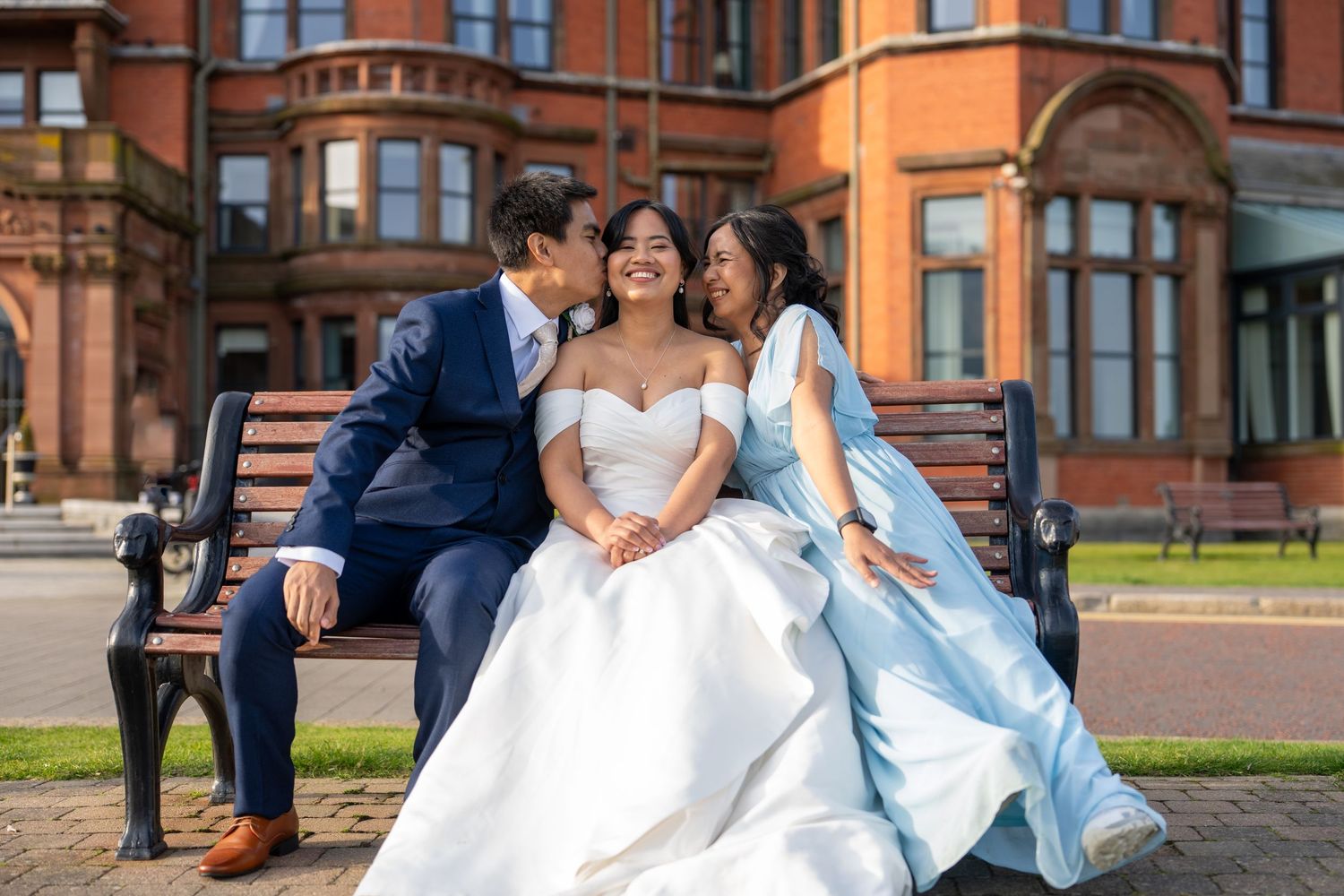 Bride in white off-shoulder gown sitting on bench with two smiling guests outside red brick building. Dave Martin Photographer Belfast