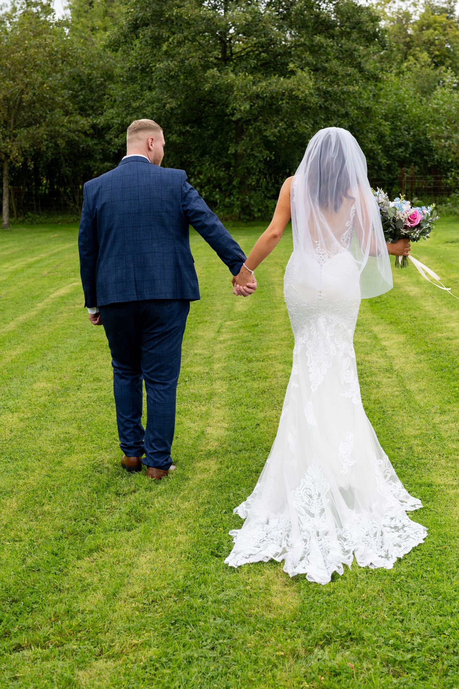 Bride and groom walking hand in hand across lush green lawn, bride in lace gown with veil. Dave Martin Photographer Northern Ireland
