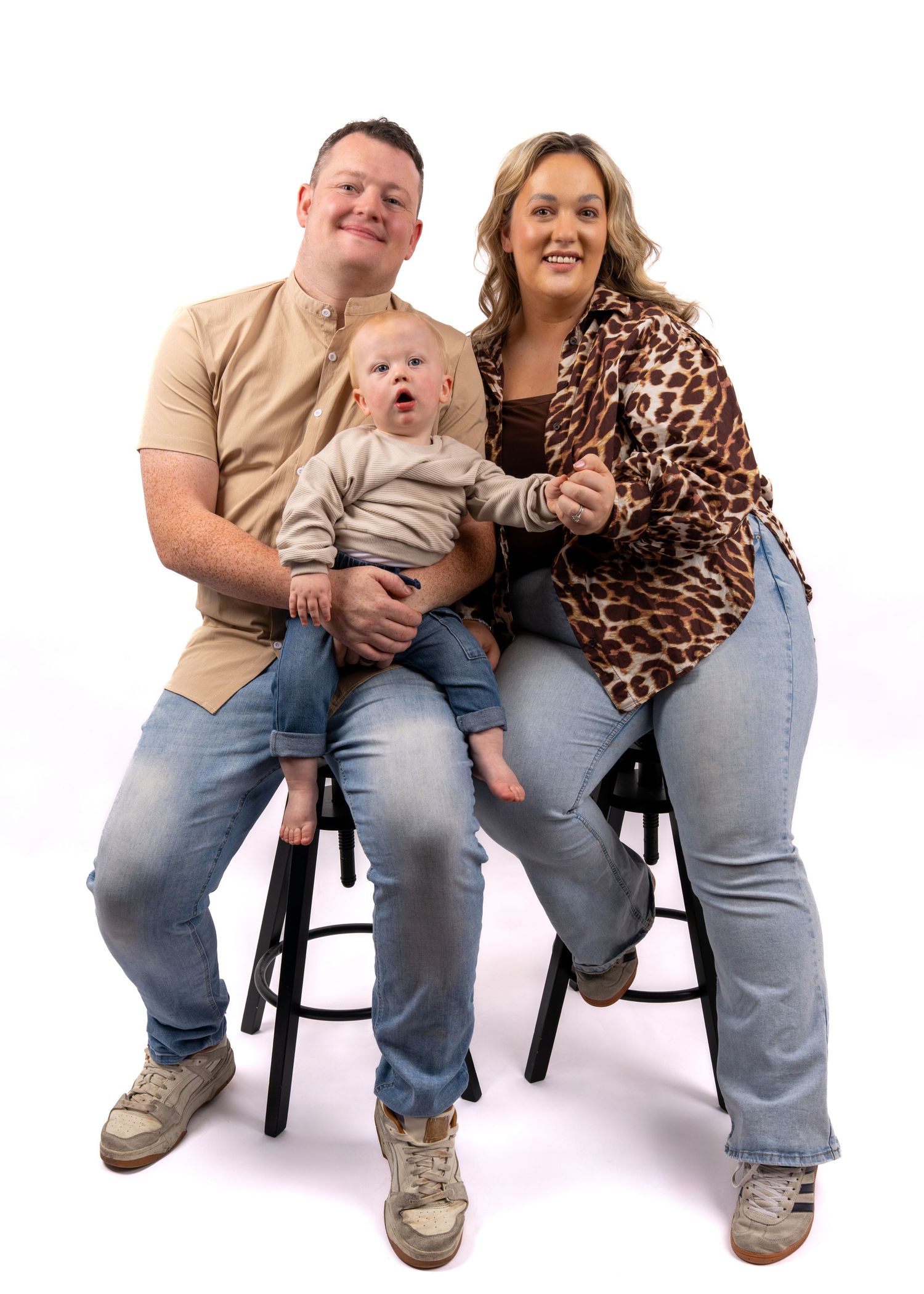 A happy family portrait with a man, woman, and baby sitting together on stools against a white background. Dave Martin Photography Studio