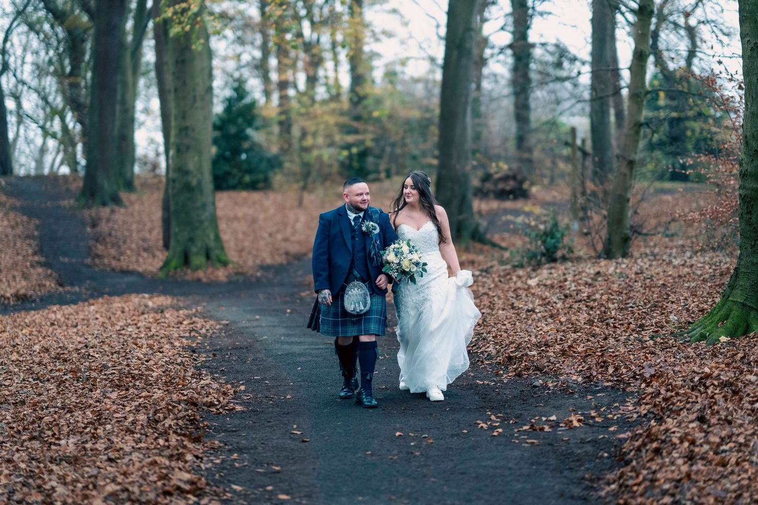 Bride and groom walking hand in hand along an autumn woodland path, groom wearing traditional Scottish kilt. By Dave Martin Photography
