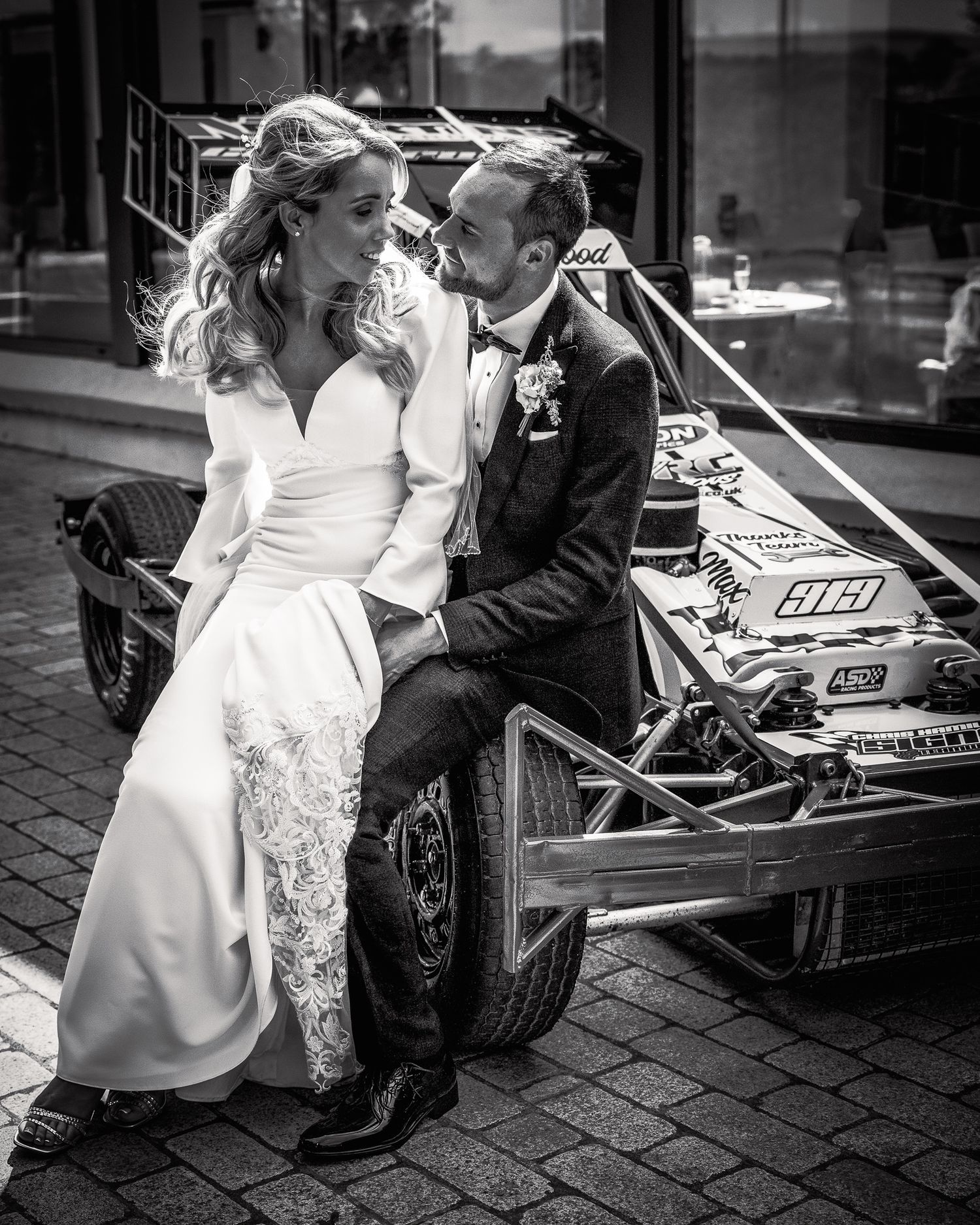 Black and white photo of a bride and groom sitting together on a vintage race car on their wedding day. Northern ireland weddings