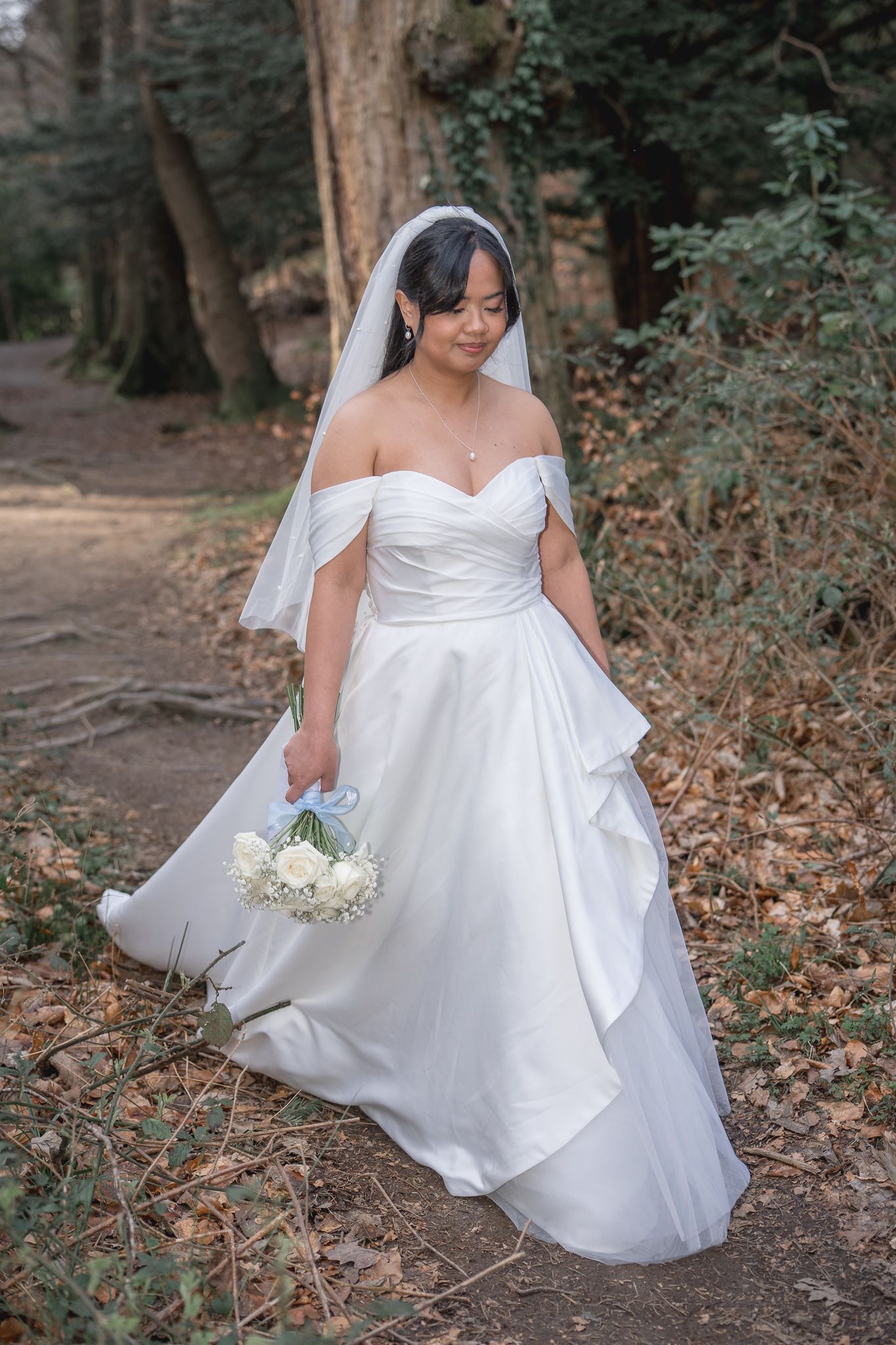 Bride in off-shoulder white gown and veil holds bouquet while walking through a wooded forest path. Dave Martin wedding Photographer NI
