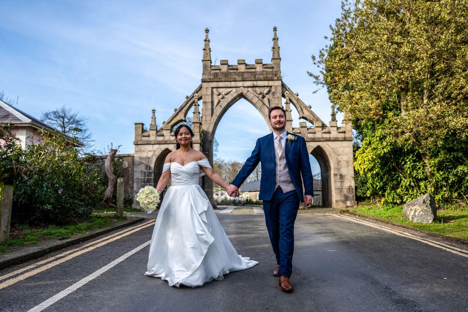 Bride and groom walking hand in hand through Gothic stone archway on sunny wedding day. Dave Martin Photographer Northern Ireland