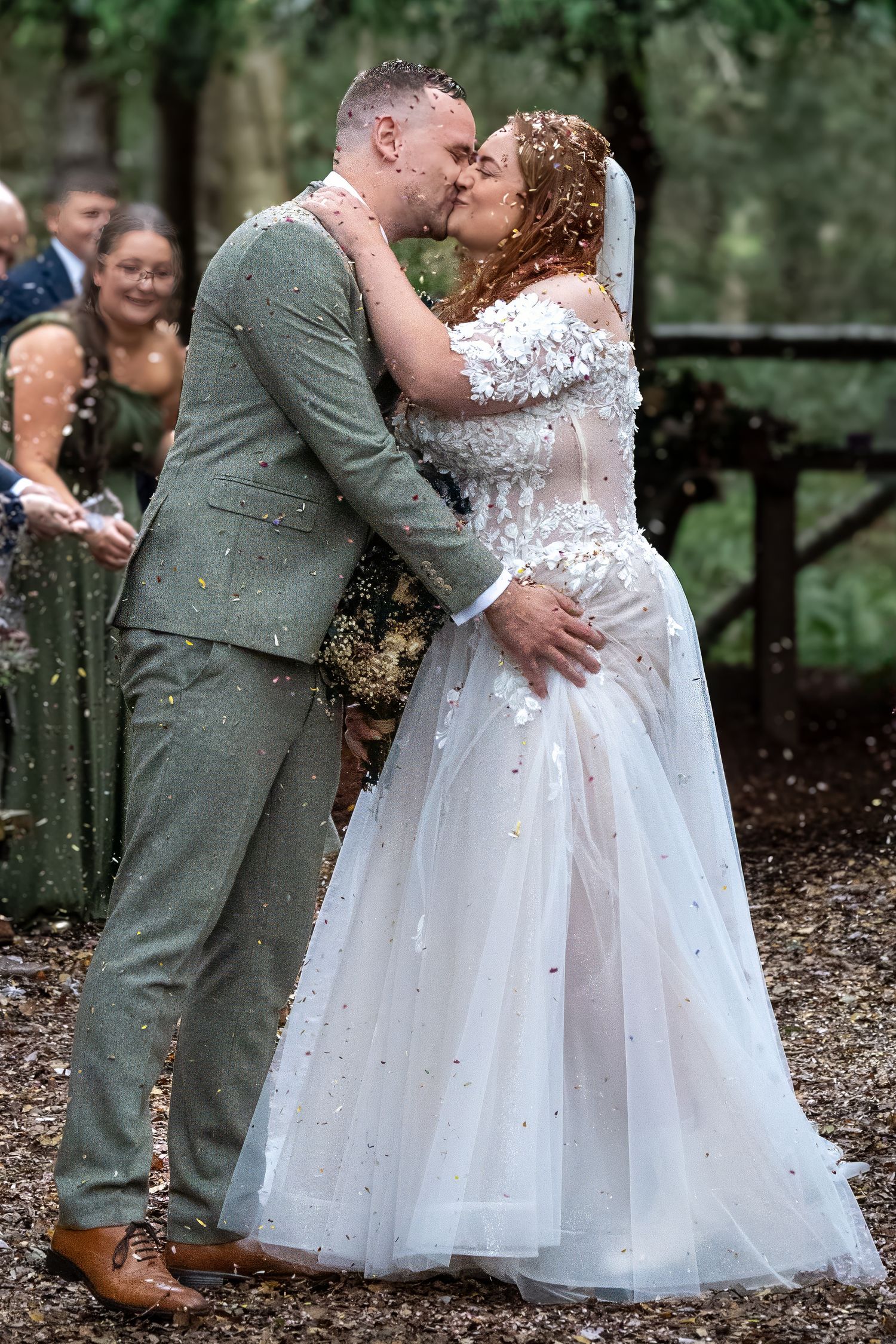 Bride and groom share first kiss as colorful confetti falls during outdoor woodland wedding ceremony. Dave Martin Photographer Northern Irel