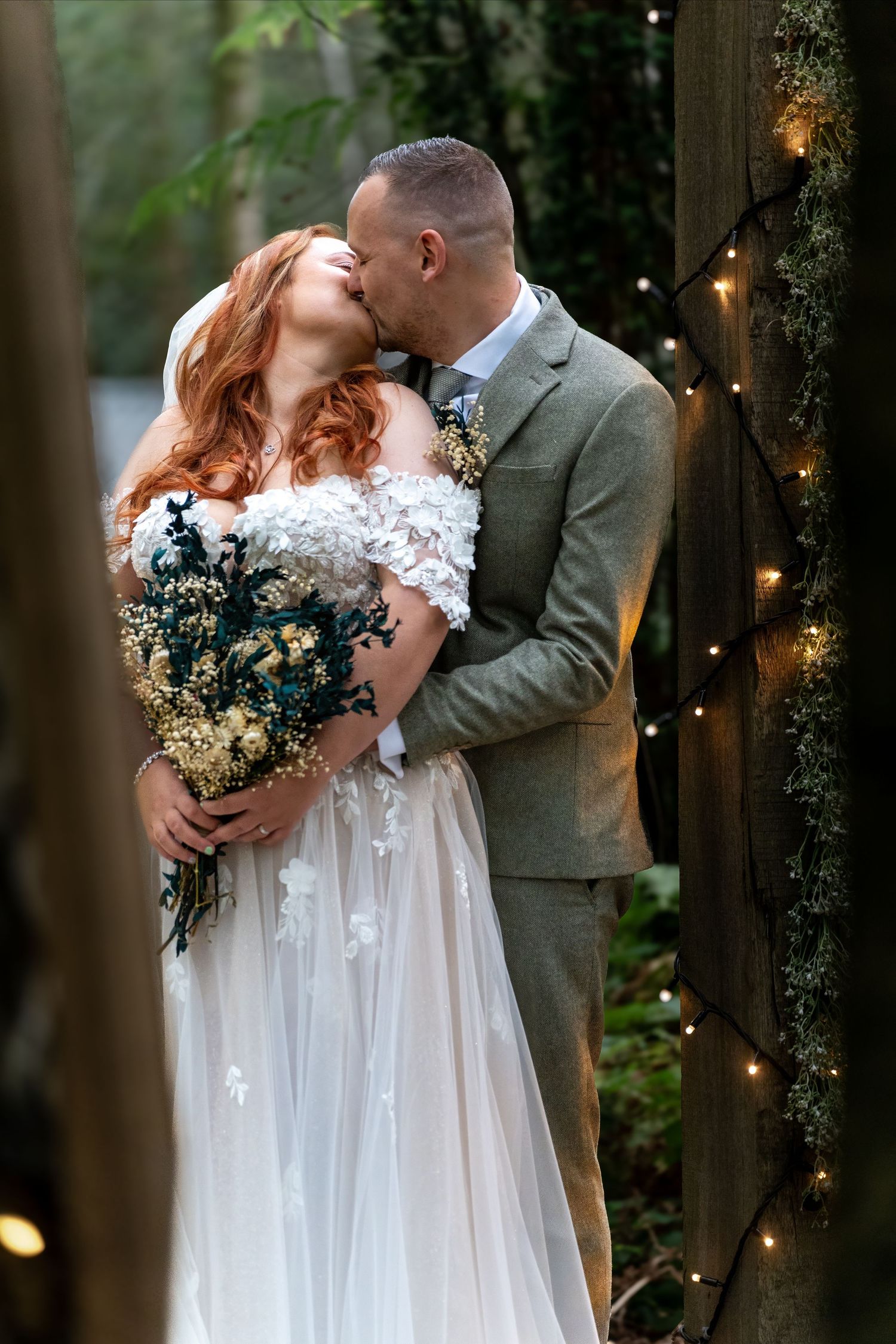 Bride with red hair holding floral bouquet shares a kiss with groom by fairy lights in forest. Dave Martin Photographer Northern Ireland