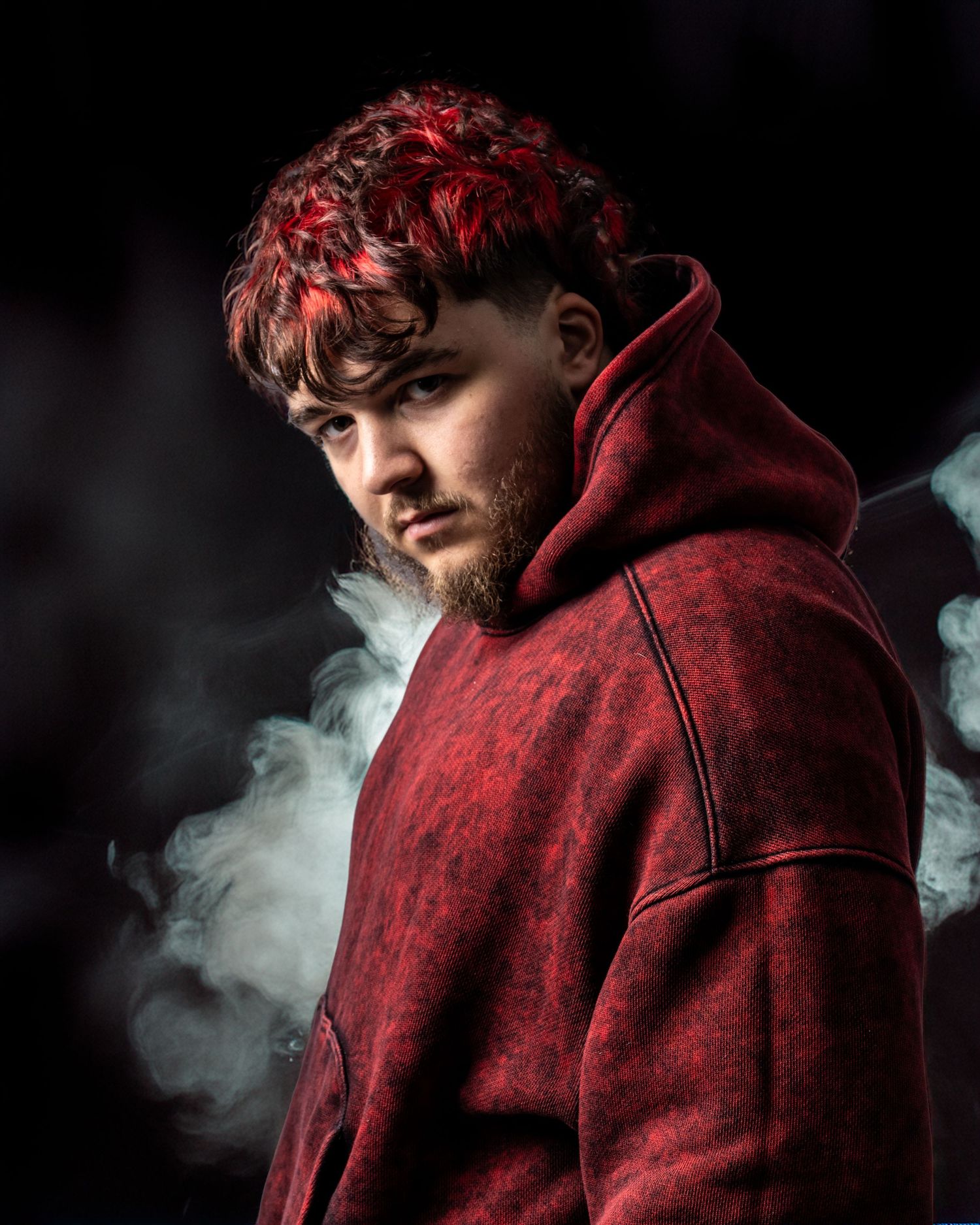 Young man with red-dyed curly hair wearing a red hoodie, surrounded by smoke against a dark background. Dave Martin Photography Studio