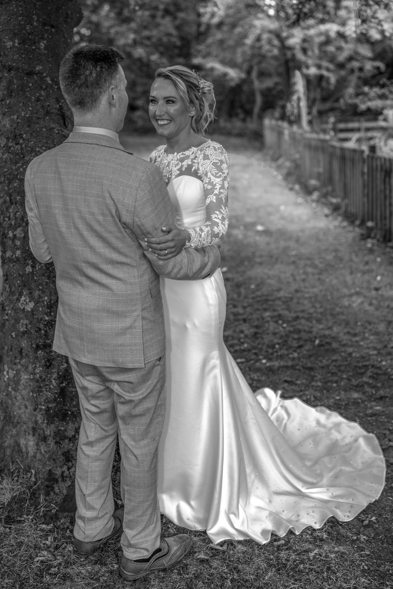 Black and white photo of a smiling bride in a satin gown embracing a groom in a suit beside a tree. Dave Martin Northern Ireland