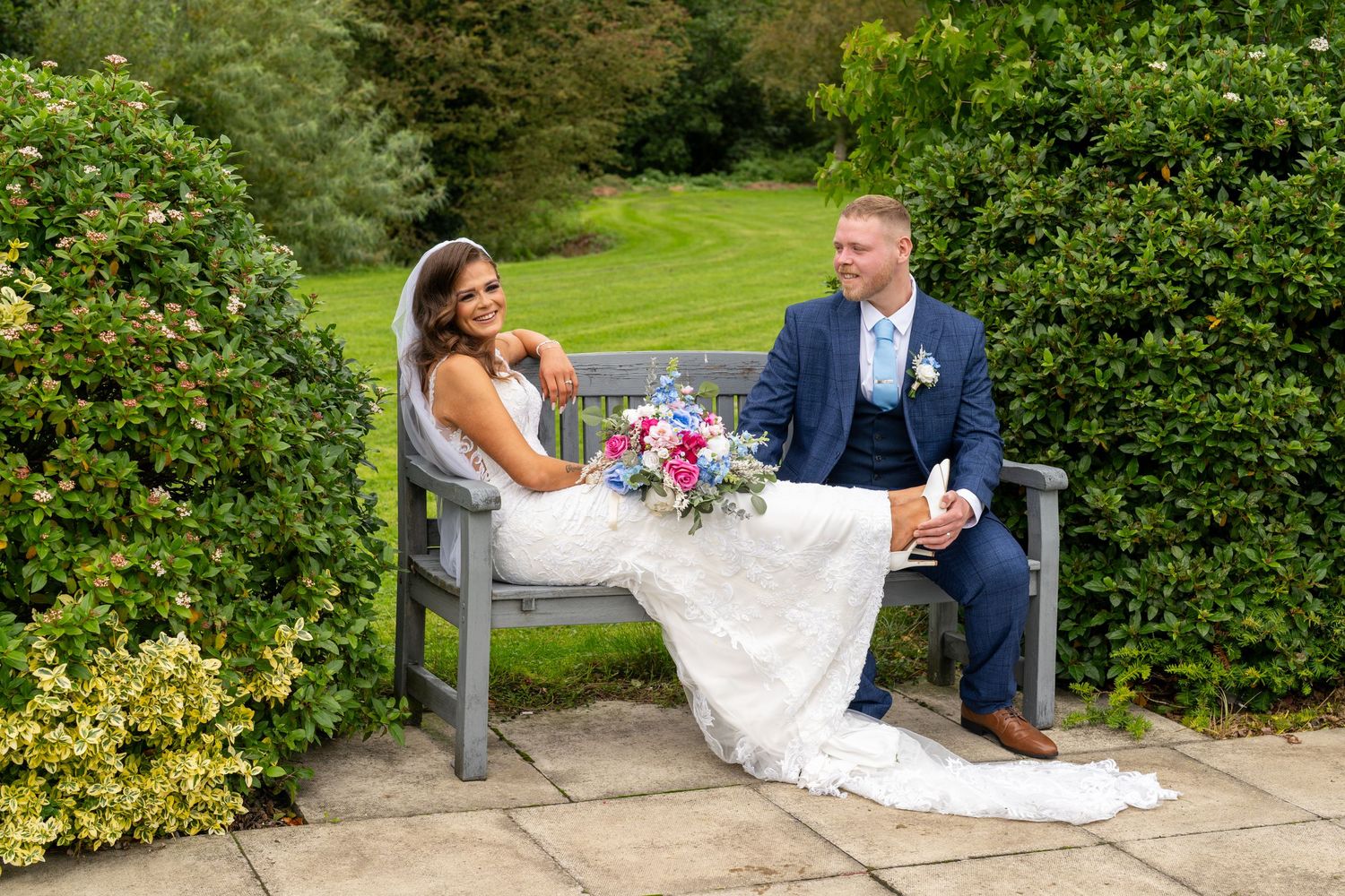 Bride and groom relaxing on garden bench surrounded by lush hedges, holding colorful bouquet. Dave Martin Photographer Northern Ireland