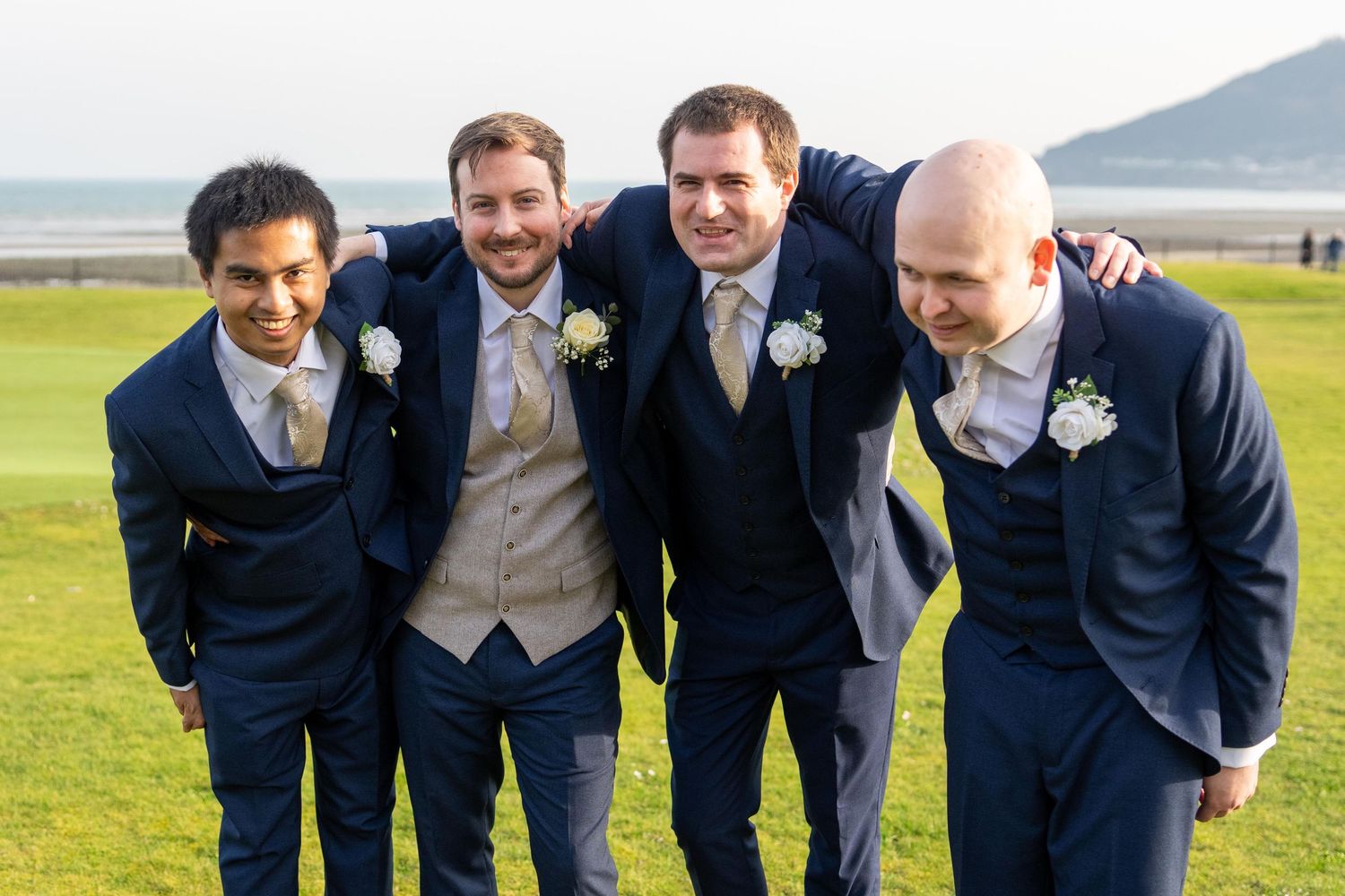 Four groomsmen in navy suits with boutonnieres laughing together outdoors at a seaside wedding venue. Slieve Donard Hotel
