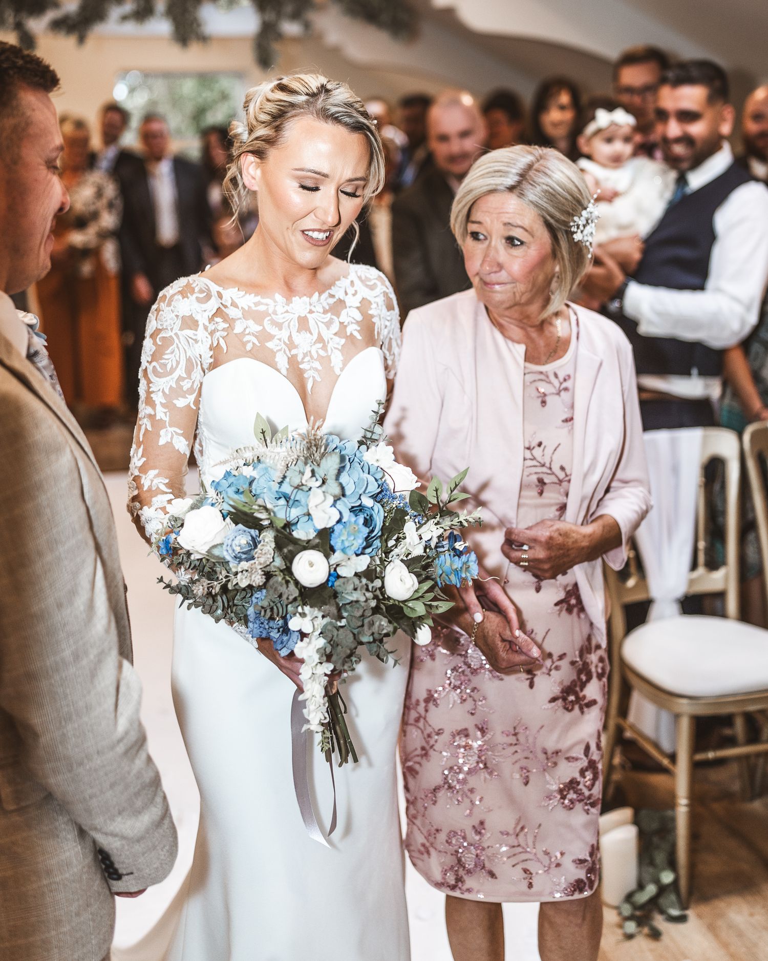 Bride in lace gown holding blue floral bouquet walks down aisle with older woman in pink dress at wedding ceremony.