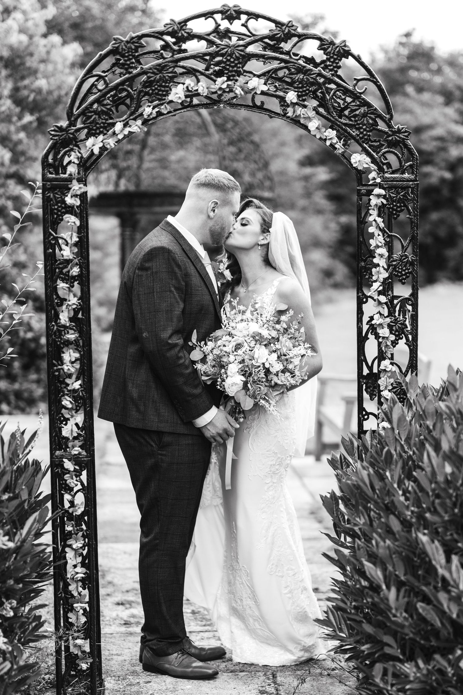Bride and groom share a romantic kiss under an ornate black iron arch in black and white. Dave Martin Photographer Northern Ireland