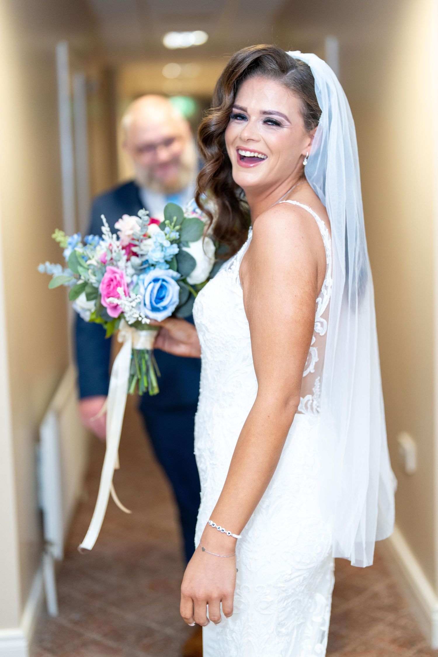 Smiling bride in white lace gown and veil holds colorful bouquet of pink and blue flowers in a hallway.