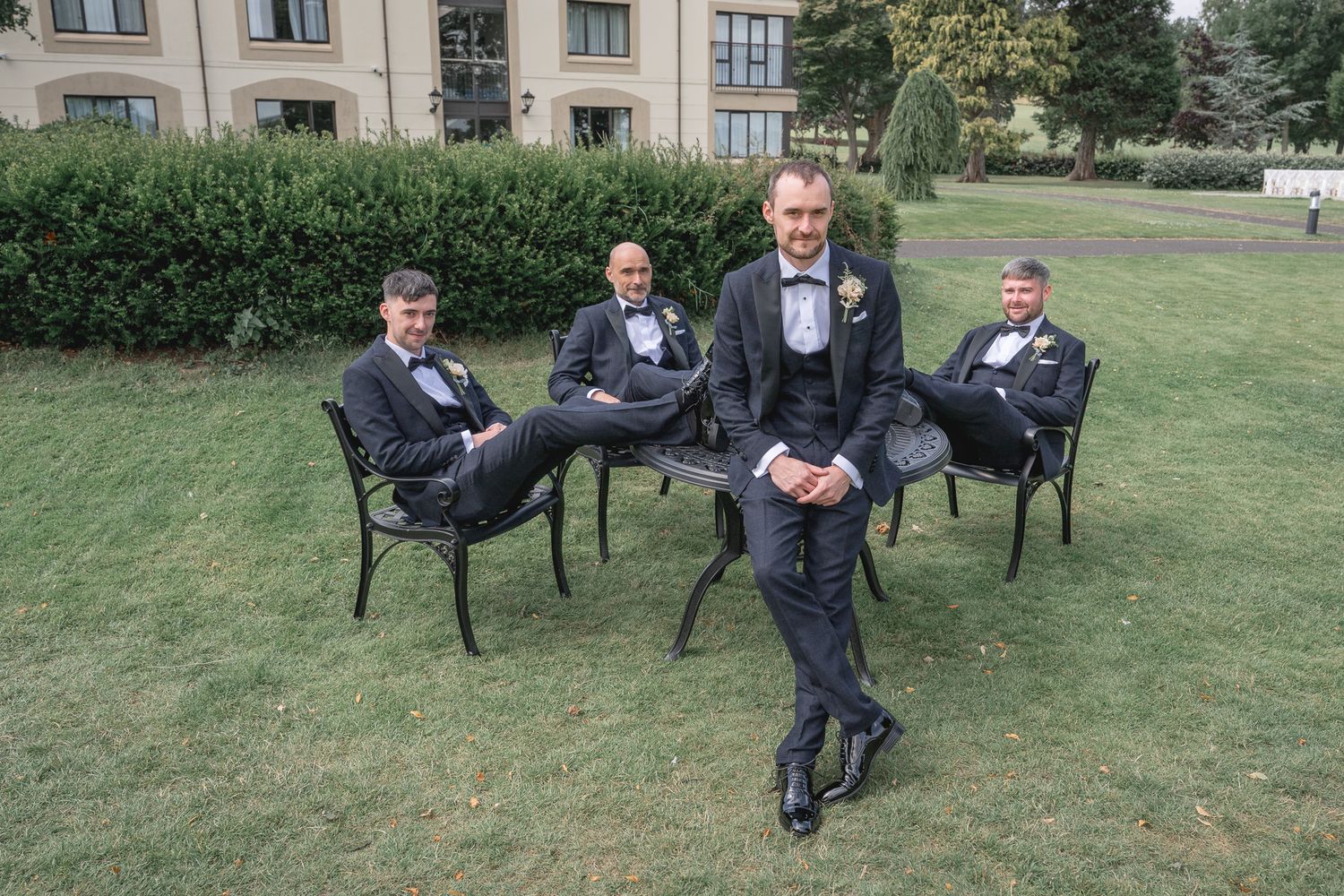 Groom and groomsmen in black tuxedos posing outdoors on green lawn near elegant building for wedding.
