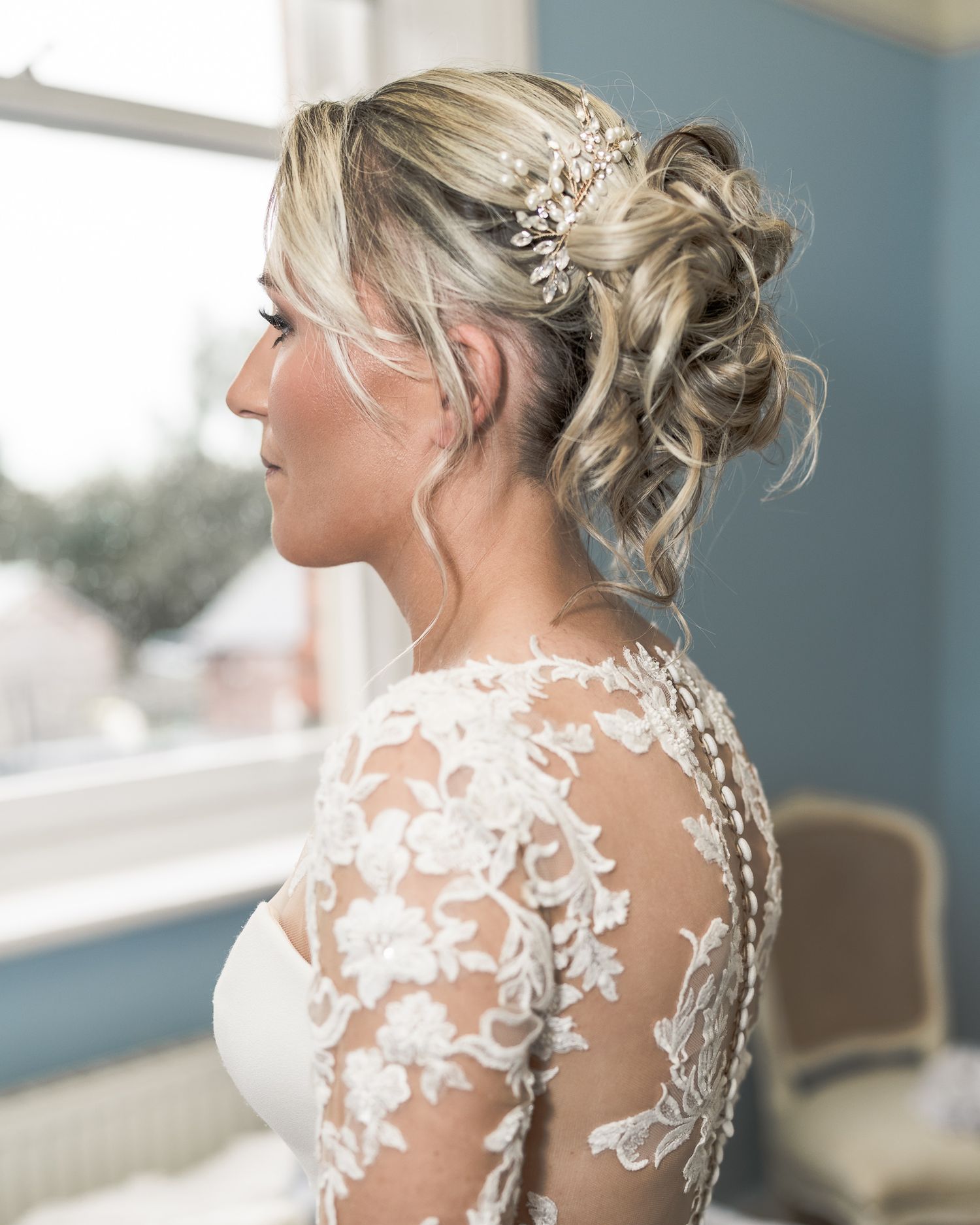 Bride in lace wedding dress with elegant updo hairstyle adorned with pearl hair comb, posing by window. Dave Martin wedding Photographer NI
