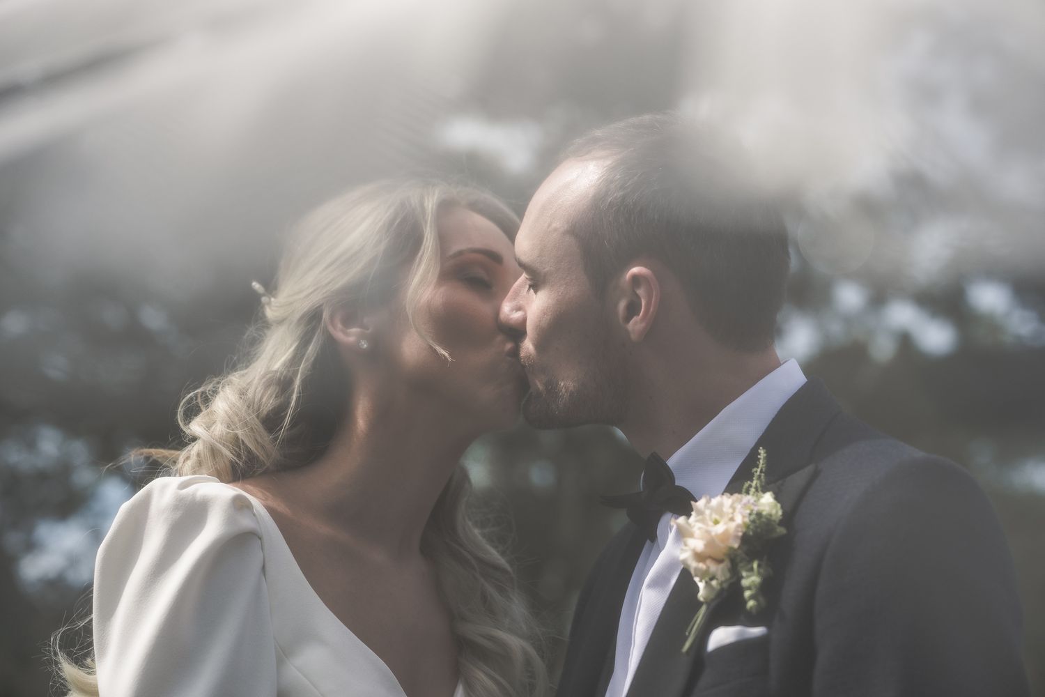 A bride and groom share a romantic kiss outdoors, bathed in soft natural light on their wedding day.  ni wedding photographer Dave Martin