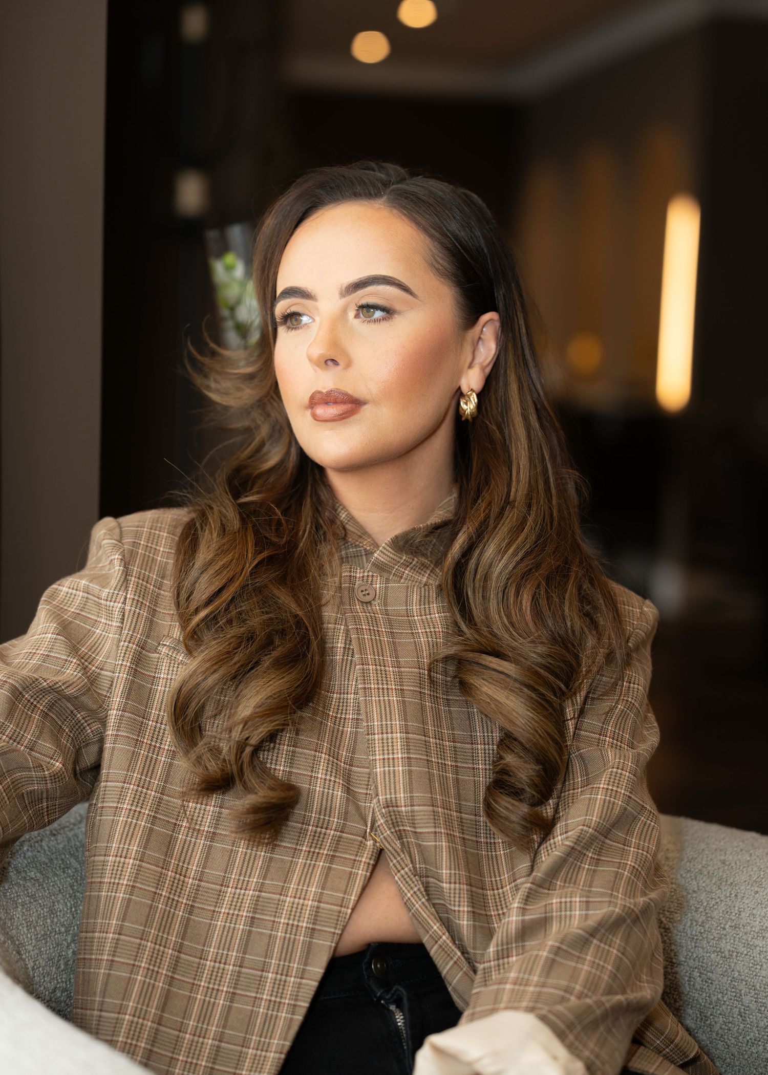 Young woman with long wavy brunette hair wearing plaid blazer seated indoors. Dave Martin Photography Northern Ireland