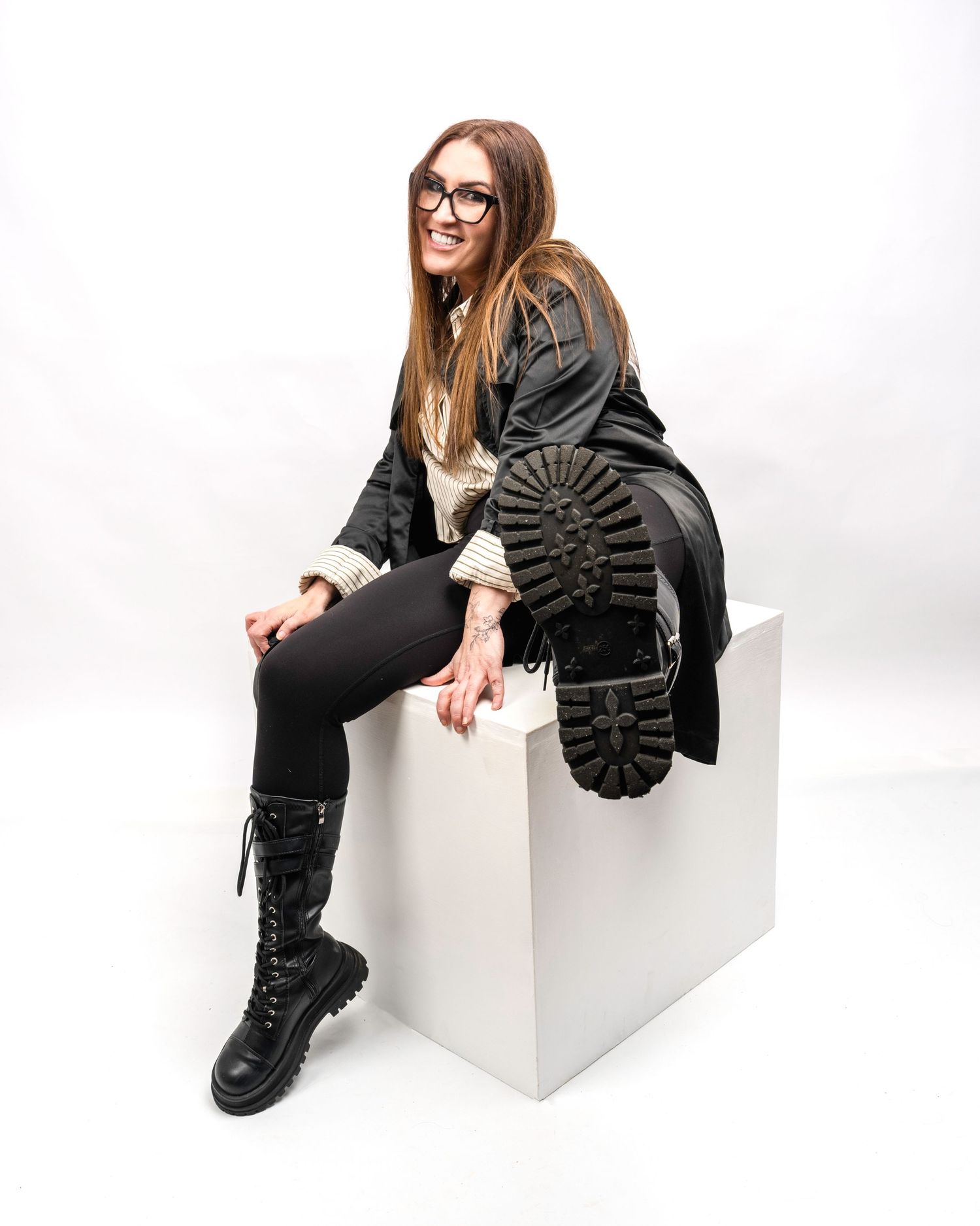 Smiling brunette woman in black coat showing boot sole while sitting on a white cube. Dave Martin Photography Studio Belfast