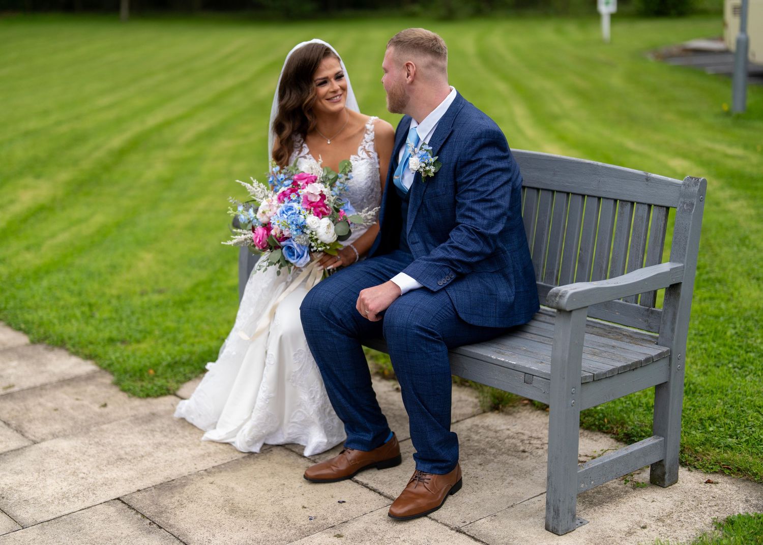 Bride and groom sitting on a gray wooden bench, smiling at each other, bride holding a colorful bouquet on a sunny day.