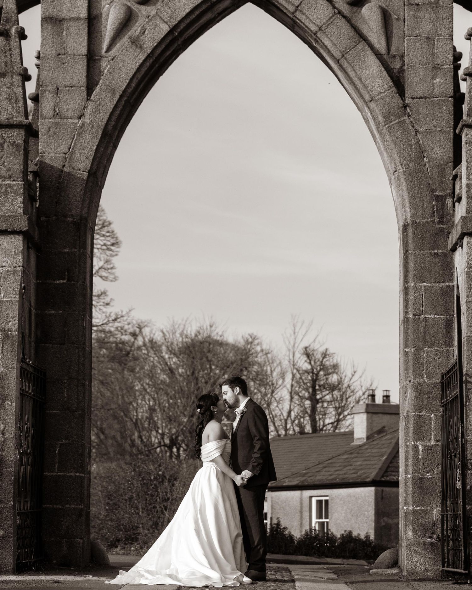 Black and white wedding photo of couple kissing under a Gothic stone arch at a historic venue.