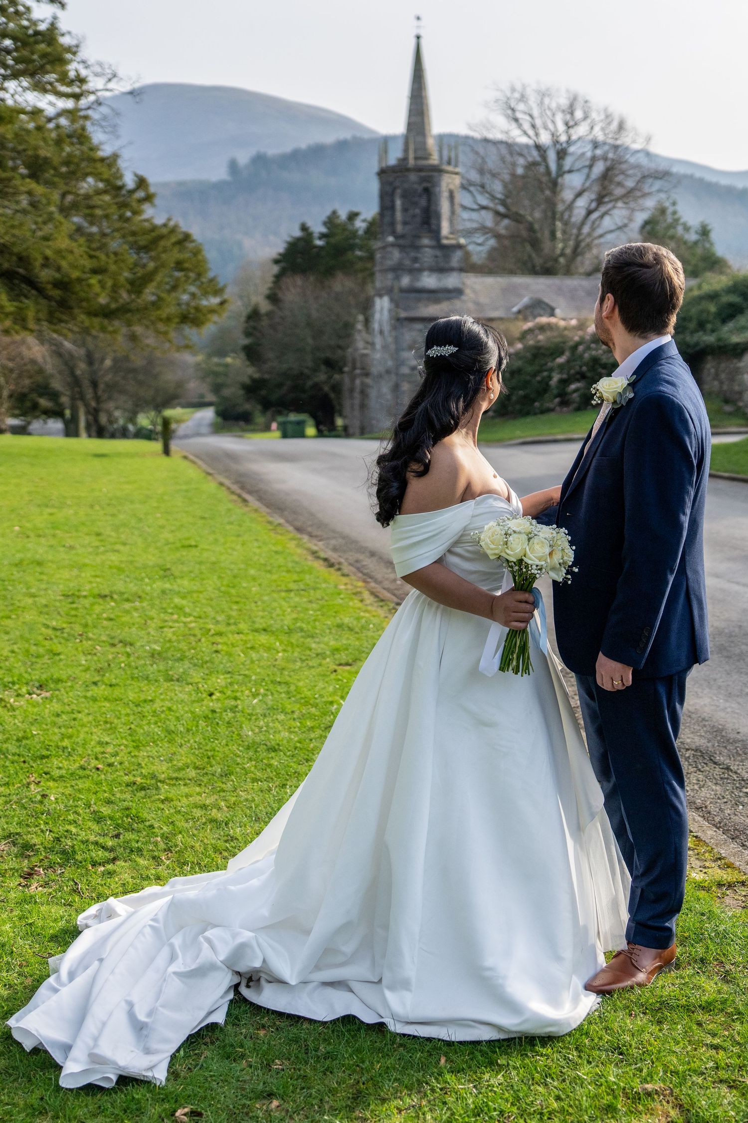 Bride and groom standing on green lawn facing historic church with mountain backdrop. Dave Martin Photographer Northern Ireland