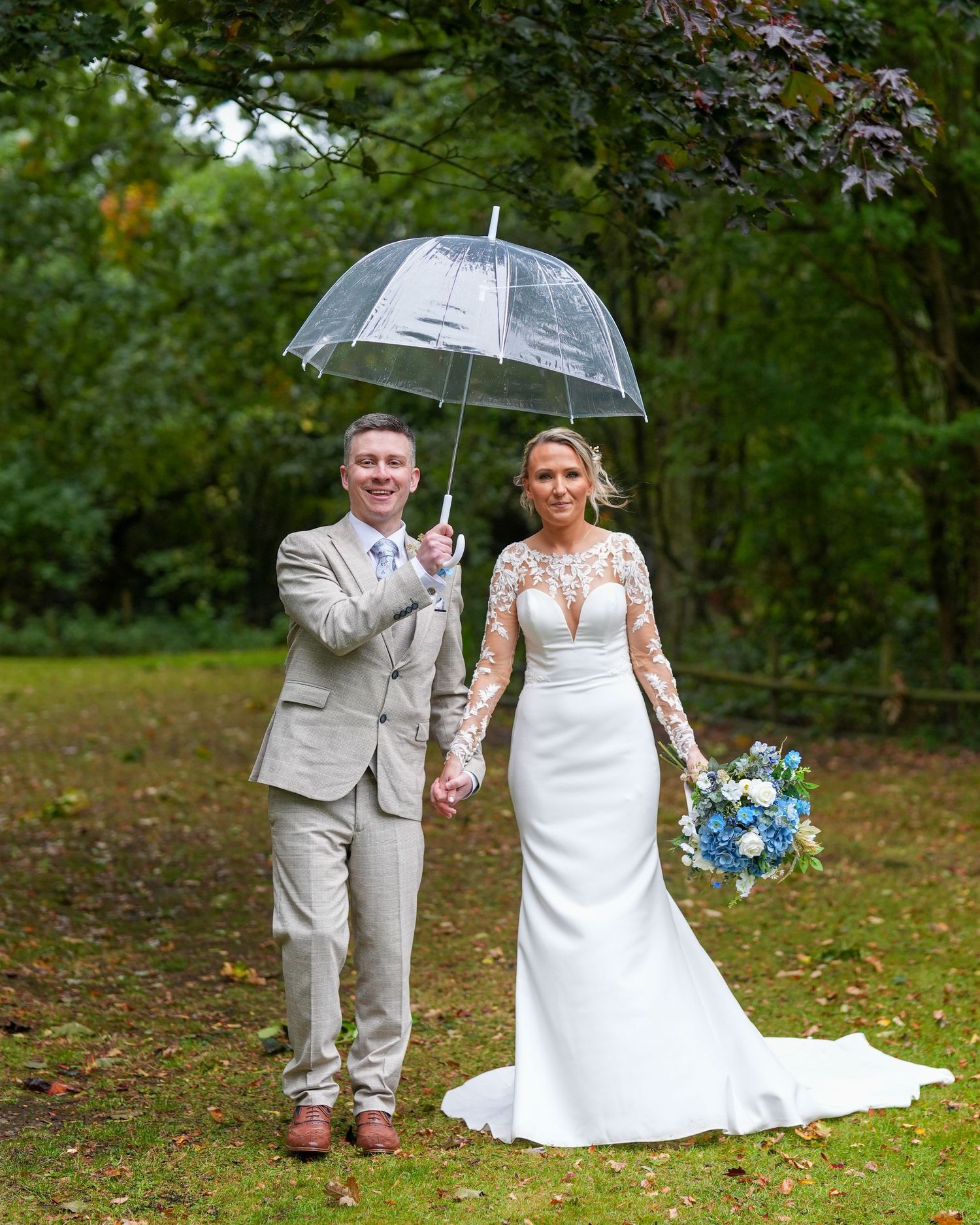 Smiling bride and groom hold a clear umbrella in a woodland garden on a rainy day. Dave Martin Photographer Northern Ireland