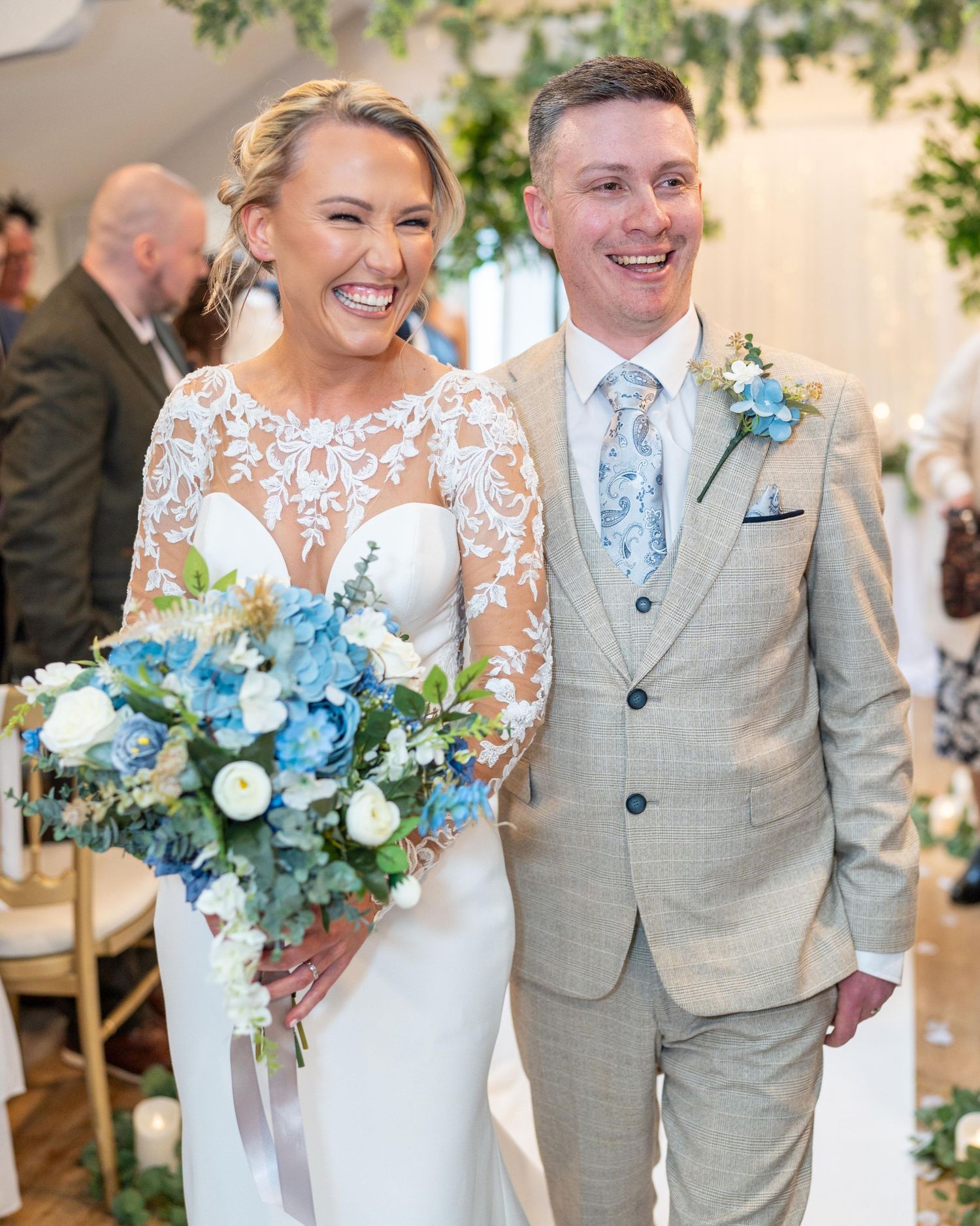 Smiling bride holding blue and white bouquet stands with groom in tan suit at elegant wedding ceremony aisle.