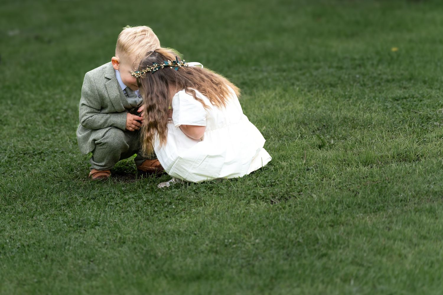Two children dressed in wedding attire crouch together on lush green grass, sharing a sweet moment. By Dave Martin Photography