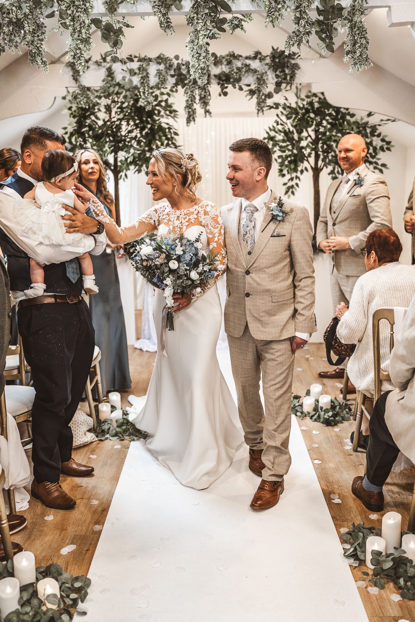 Bride and groom walk down the aisle smiling, surrounded by guests, greenery, and candles in an elegant wedding venue.
