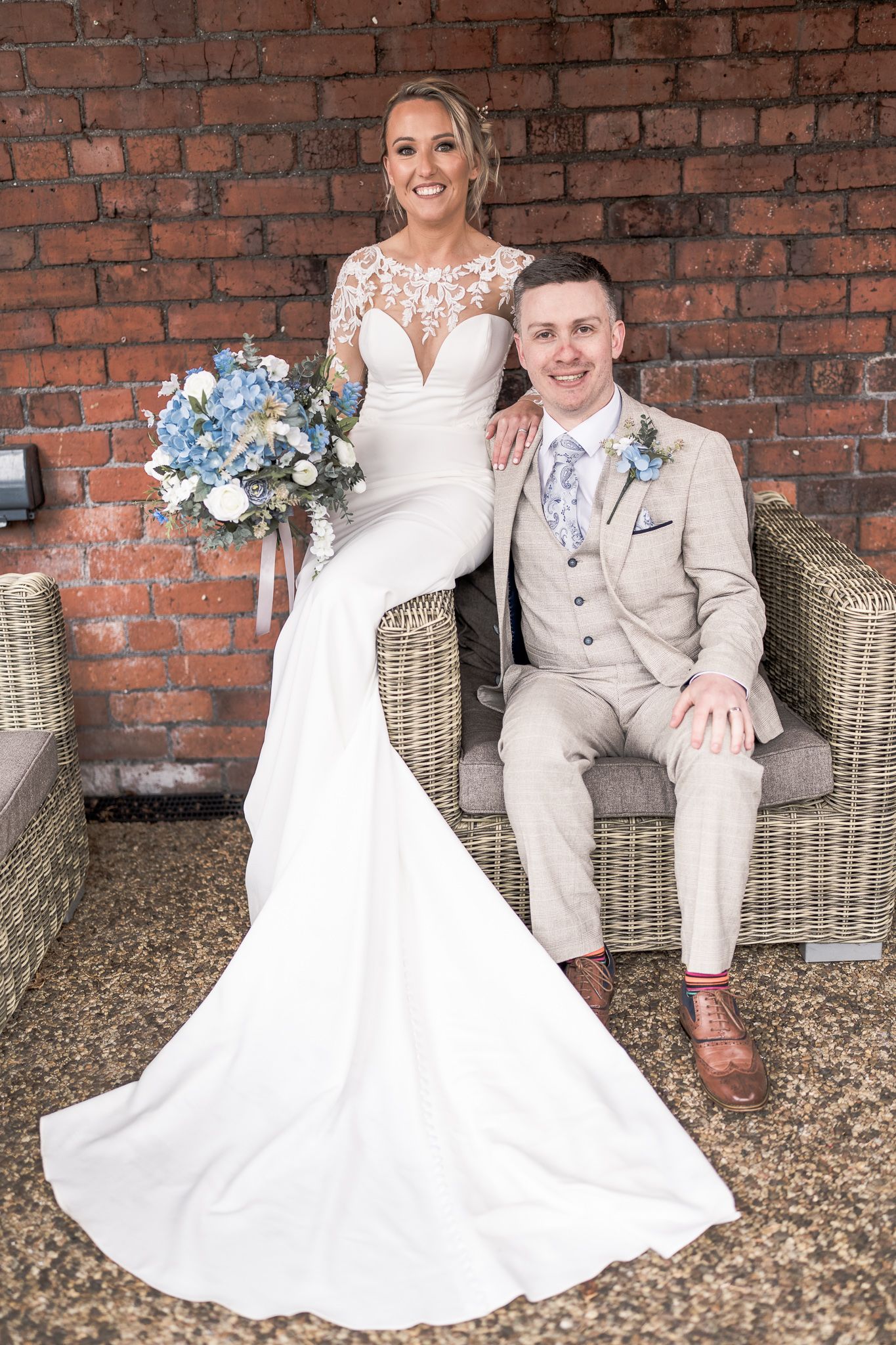Bride in white lace gown holding blue bouquet poses with groom in beige suit on wicker chair against brick wall. Dave Martin wedding Photo