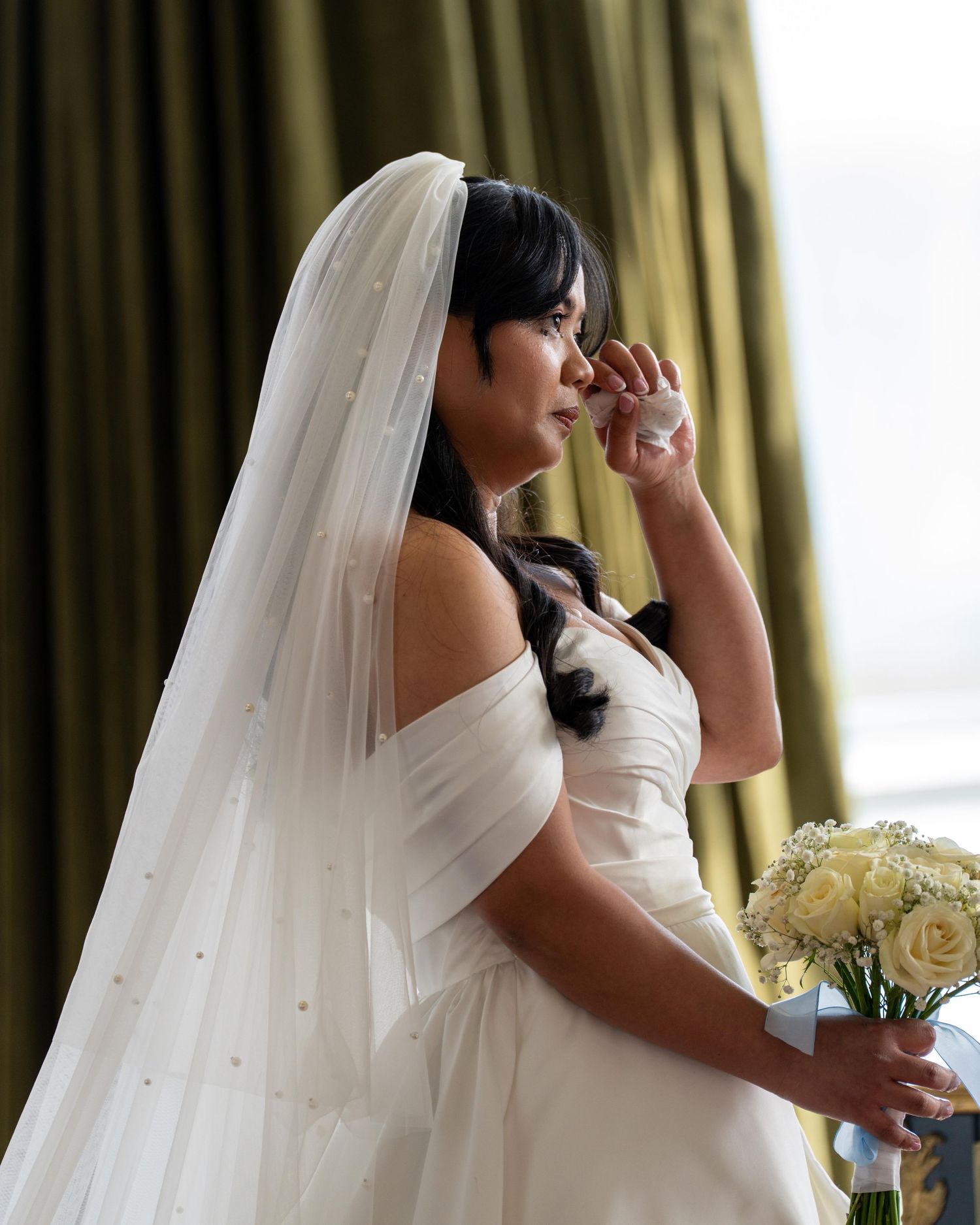 Emotional bride in white off-shoulder gown and veil wipes tear, holding white rose bouquet near draped curtains. Dave Martin Weddings
