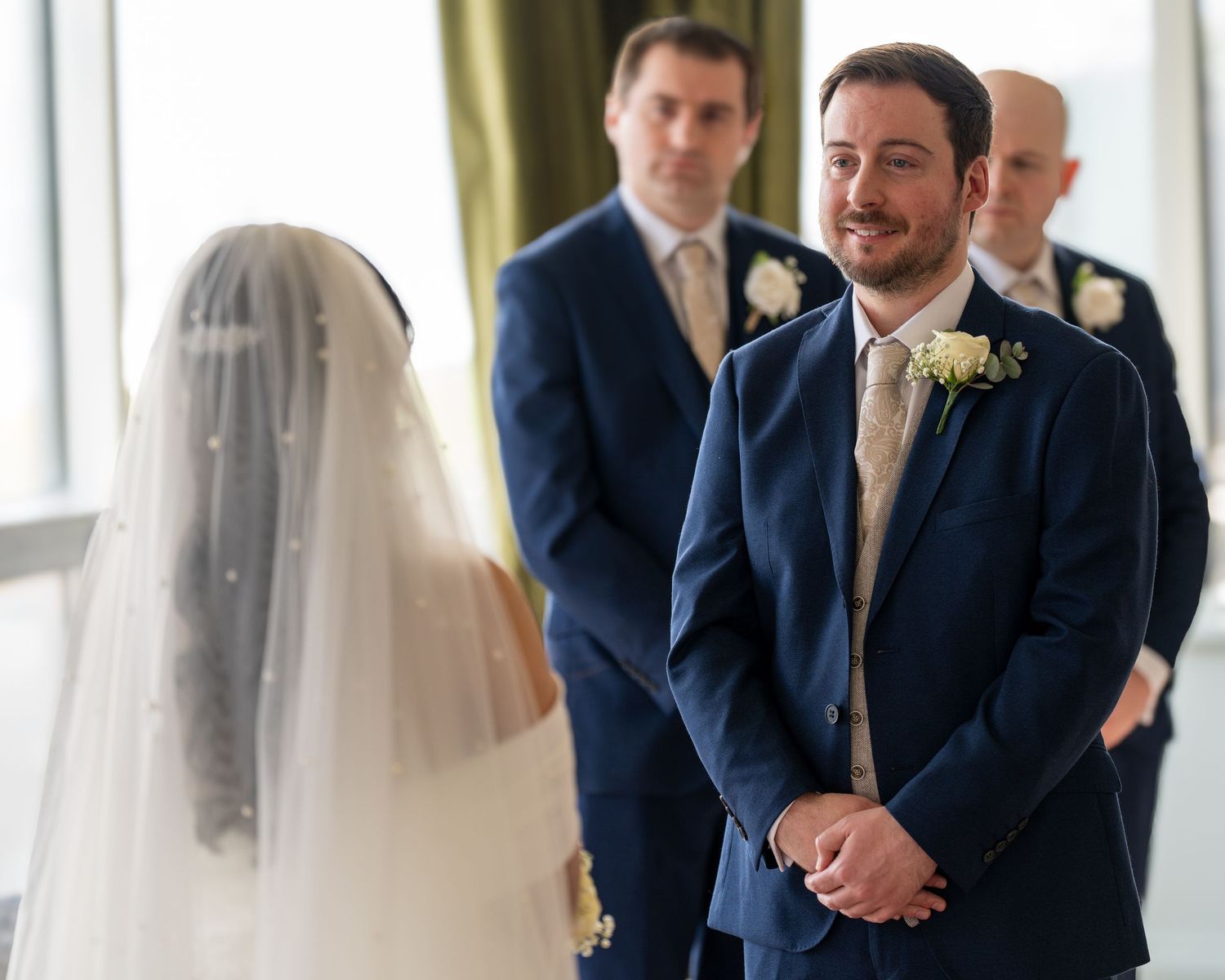 Groom in navy suit smiling warmly at veiled bride during indoor wedding ceremony. Dave Martin Photographer Northern Ireland