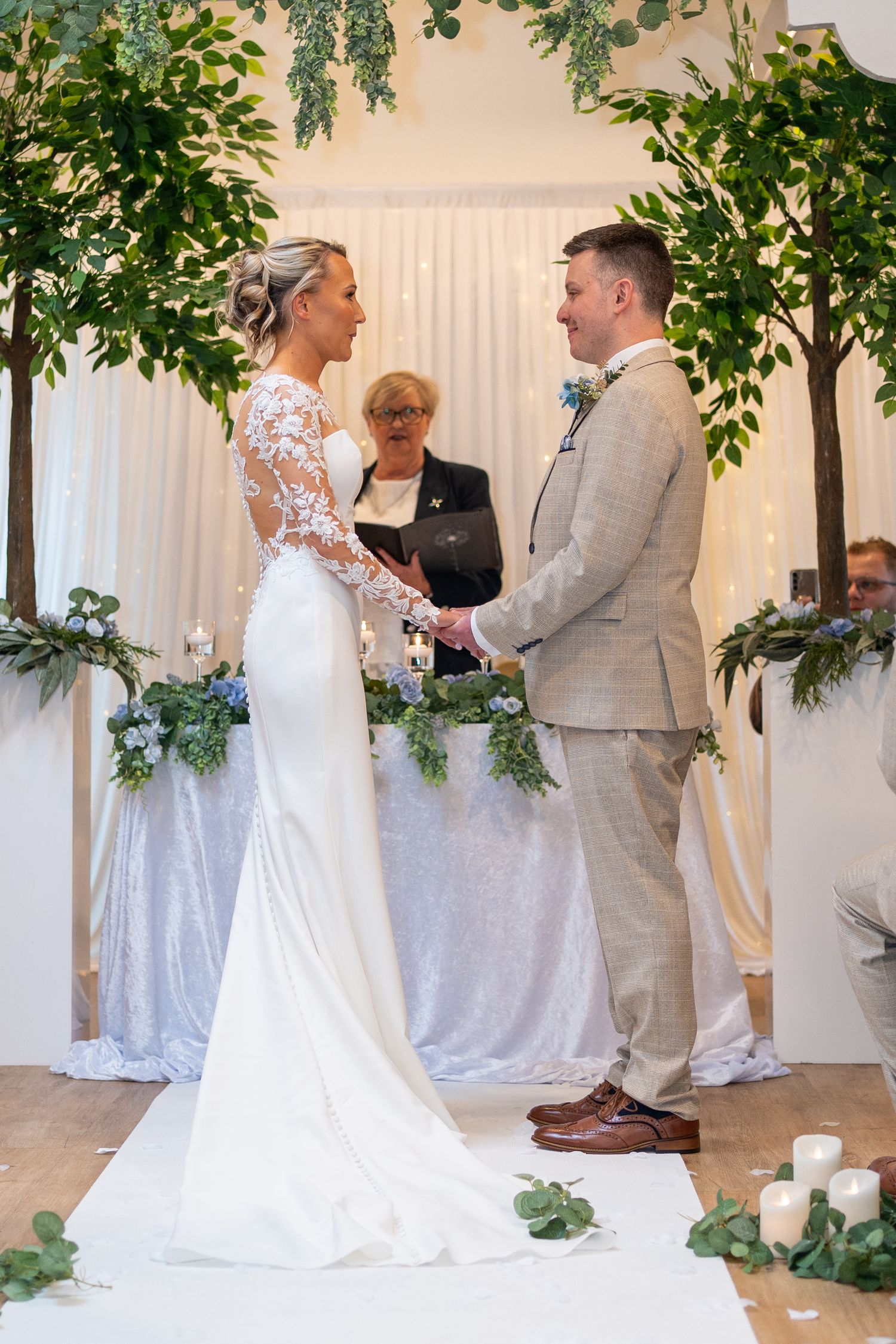 Bride and groom exchange vows at floral arch altar, officiant behind them, indoor wedding ceremony.