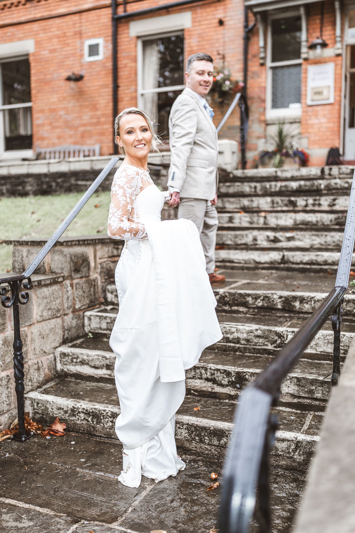 Bride in white lace gown holds dress train on stone steps while groom in gray suit stands behind her smiling. Dave Martin wedding Photograph
