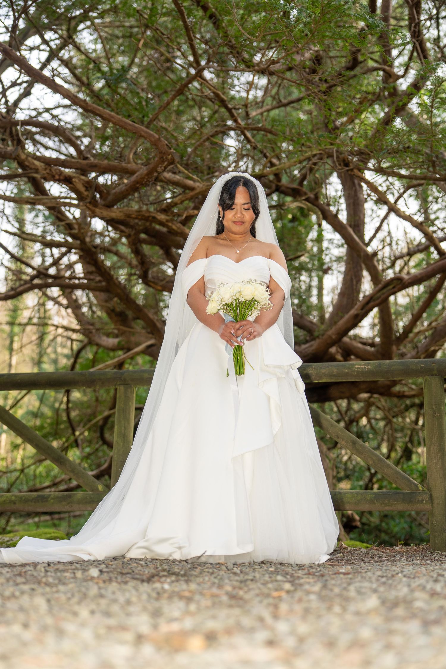 Bride in off-shoulder white ball gown and veil holds bouquet before rustic fence and trees.