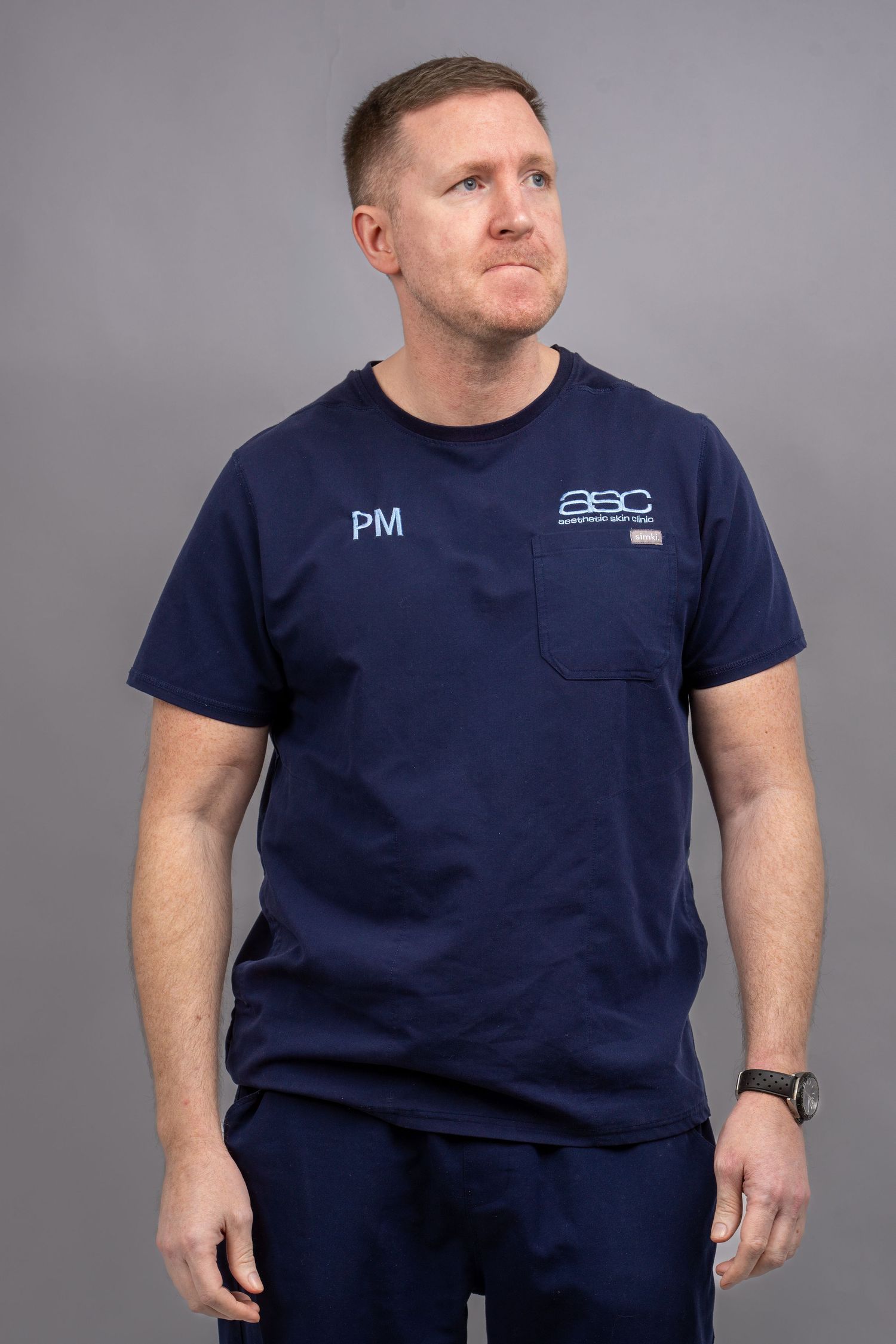 Man wearing navy blue scrubs with PM initials and ASC logo embroidered on chest, standing against gray background. Dave Martin Photography
