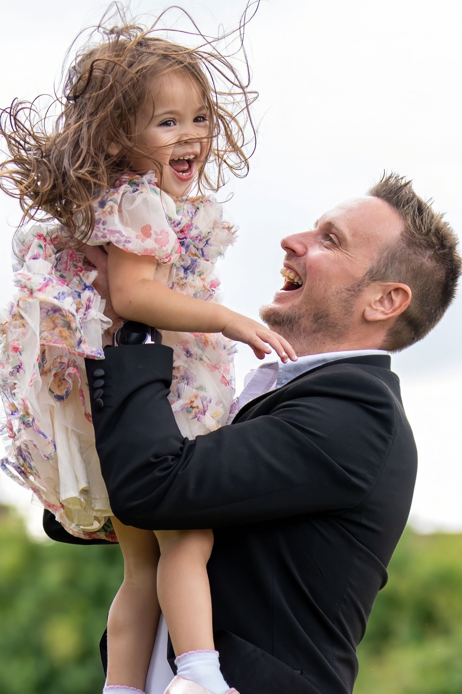 Joyful father in black suit lifts laughing little girl in floral dress outdoors. Dave Martin Photographer Northern Ireland