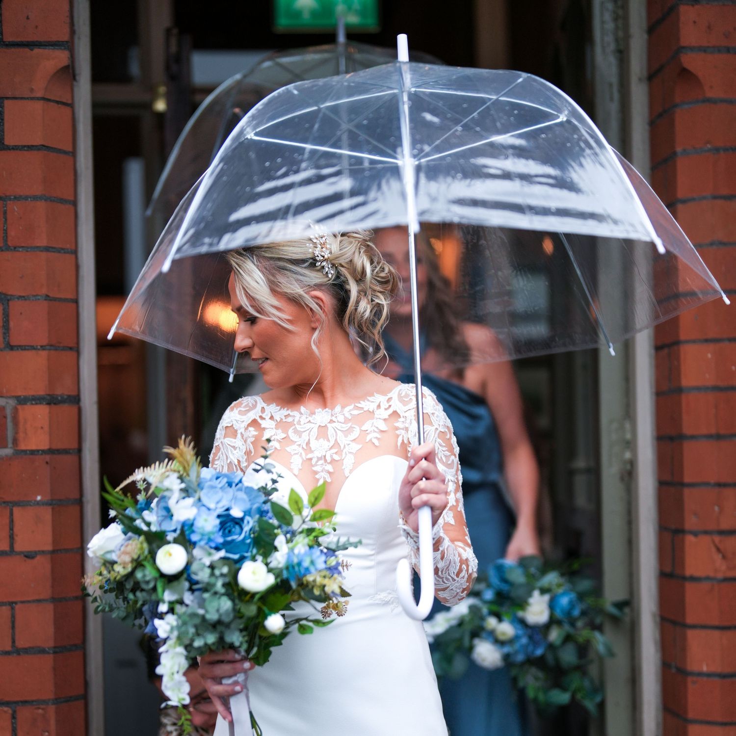 Bride with lace gown and blue hydrangea bouquet shelters under clear umbrella at venue entrance. Dave Martin Photographer Northern Ireland