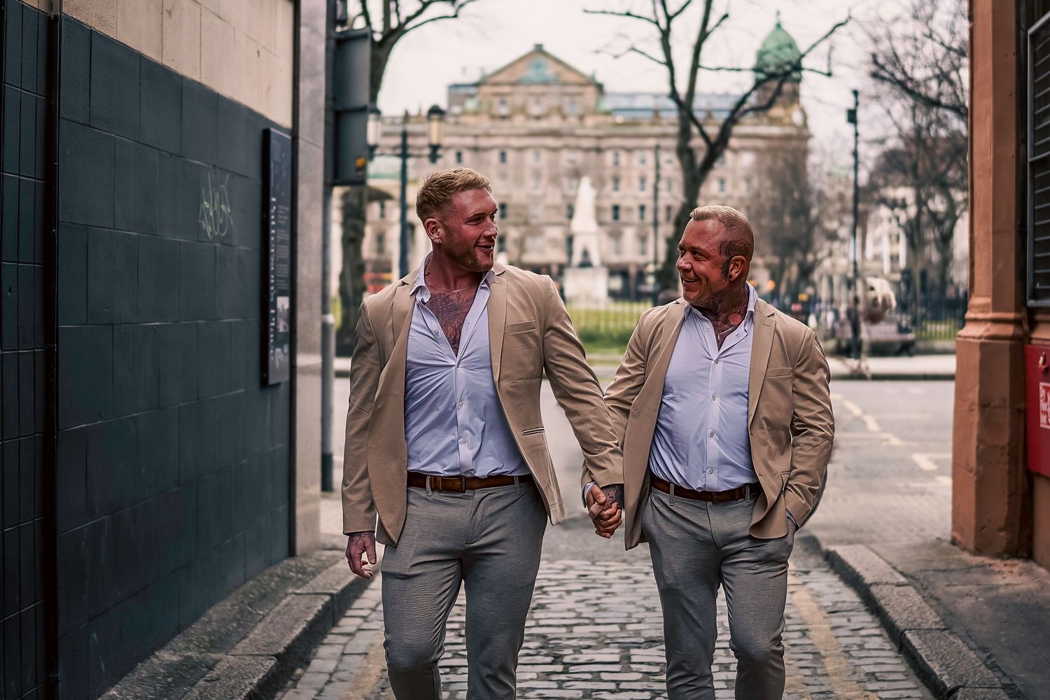 Two men in matching beige blazers and grey trousers hold hands while walking and smiling at each other on a city street. By Dave Martin Phot