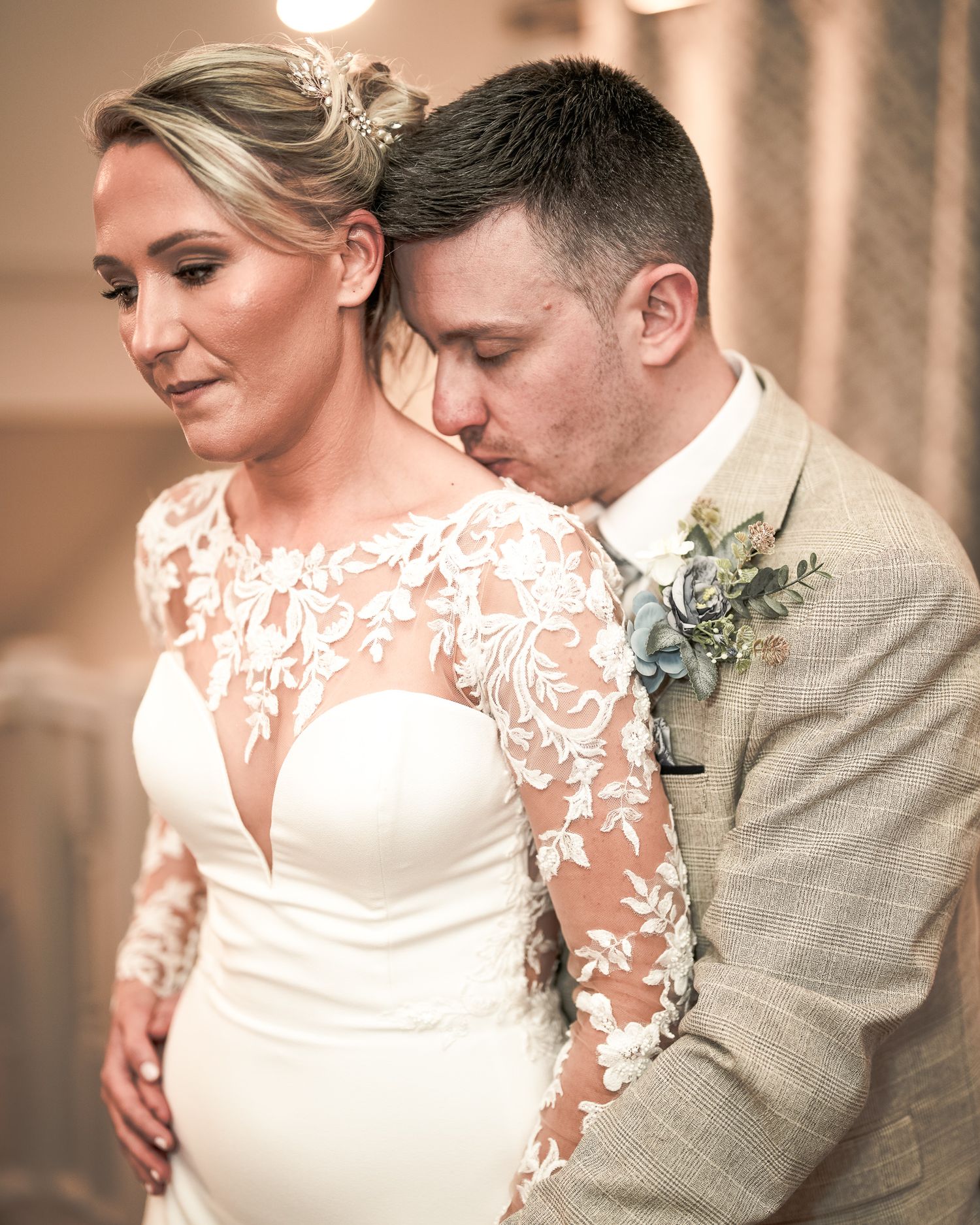 Groom in tan suit kissing bride's cheek as she wears lace long-sleeve wedding gown at elegant indoor ceremony. Dave Martin wedding Photos
