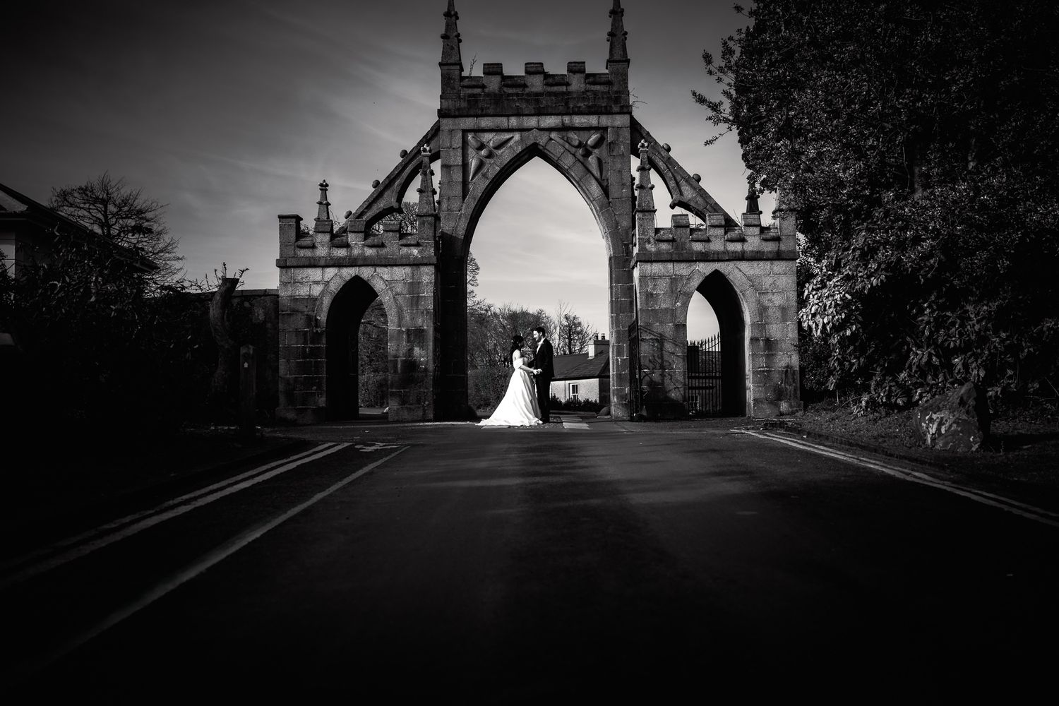 Black and white wedding photo of bride and groom standing beneath a grand Gothic stone archway on a quiet road.