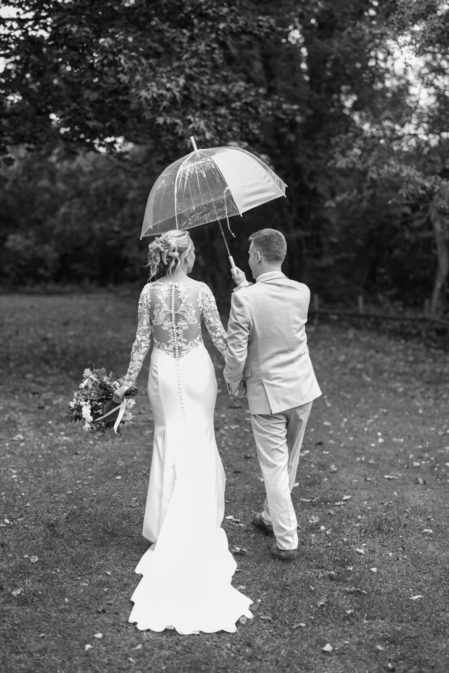 Black and white photo of bride and groom walking hand in hand under a clear umbrella on their rainy wedding day.