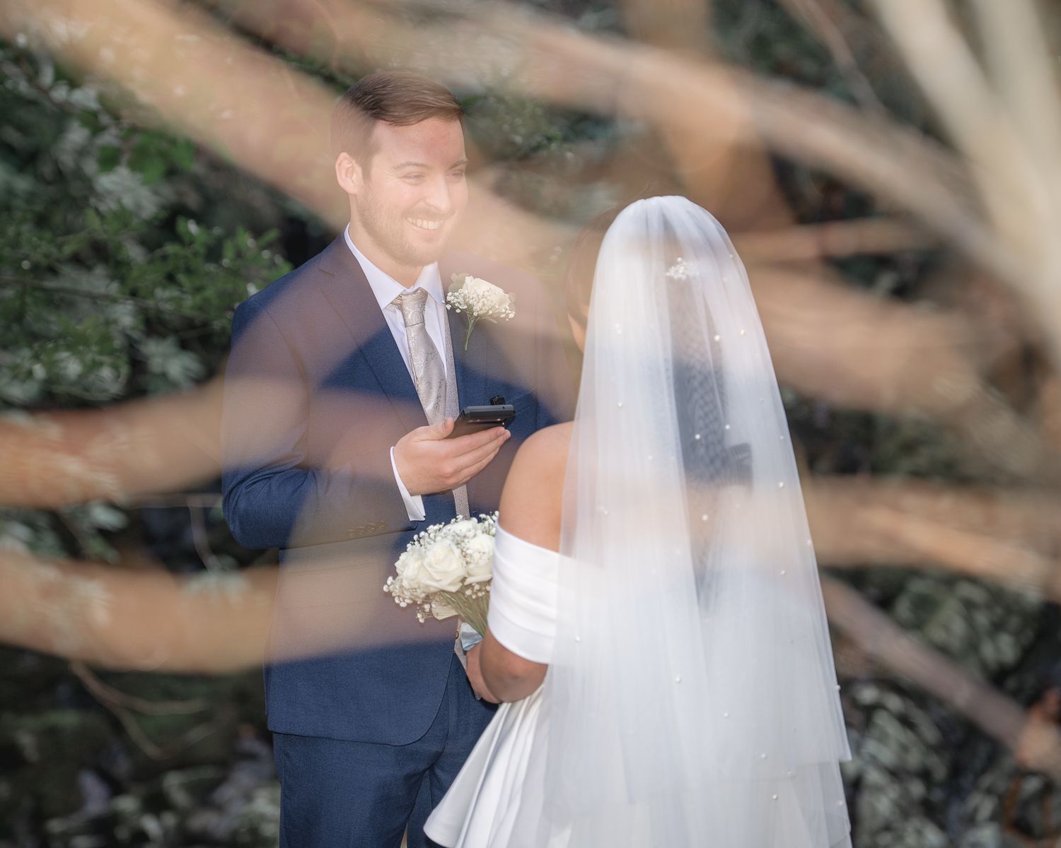 Bride in white gown and veil holds bouquet facing groom in navy suit during outdoor wedding ceremony. Dave Martin wedding Photographer NI