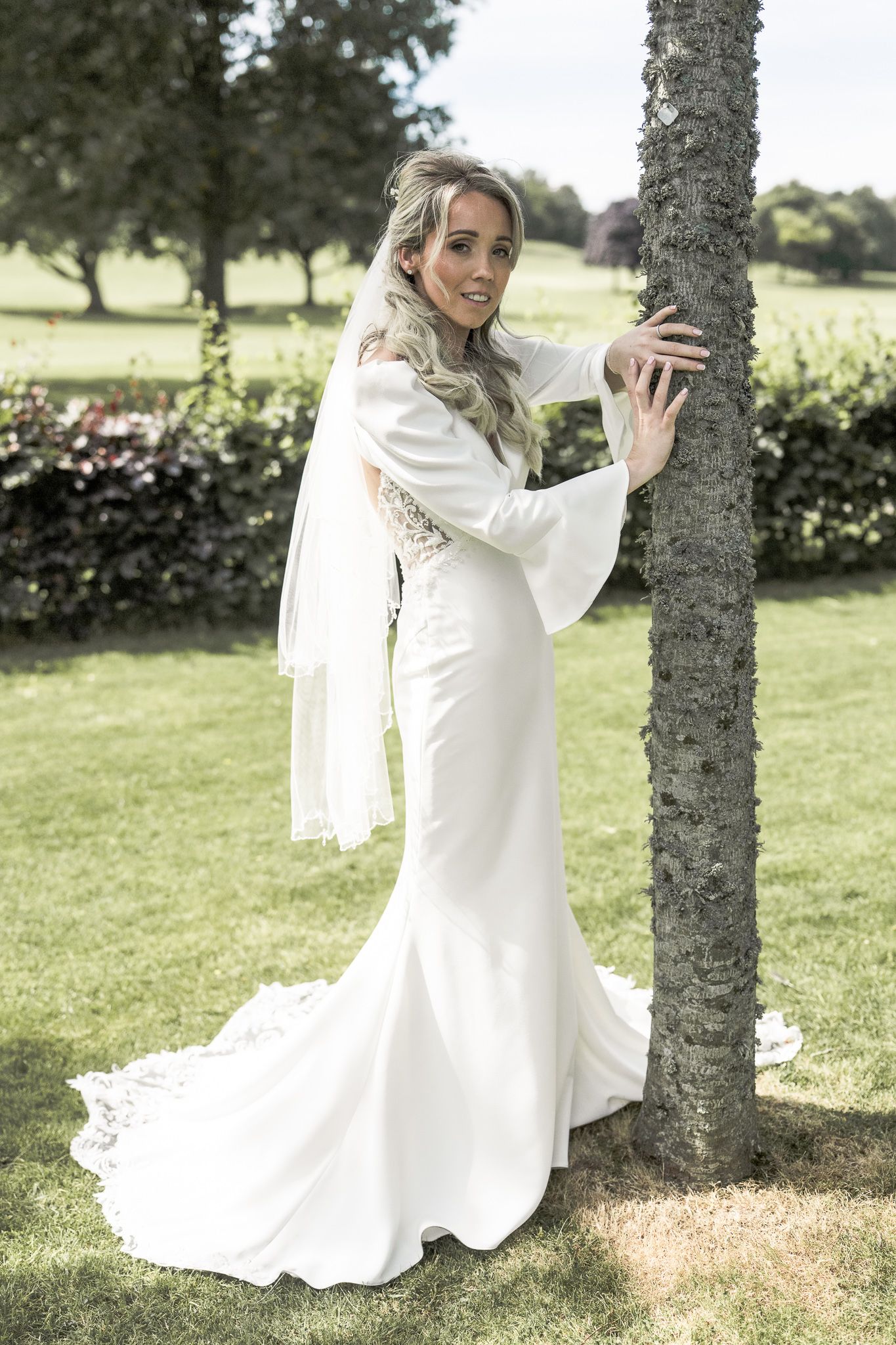 Bride in elegant white mermaid wedding gown with bell sleeves and veil, posing by a tree in a lush garden.  ni wedding photographer