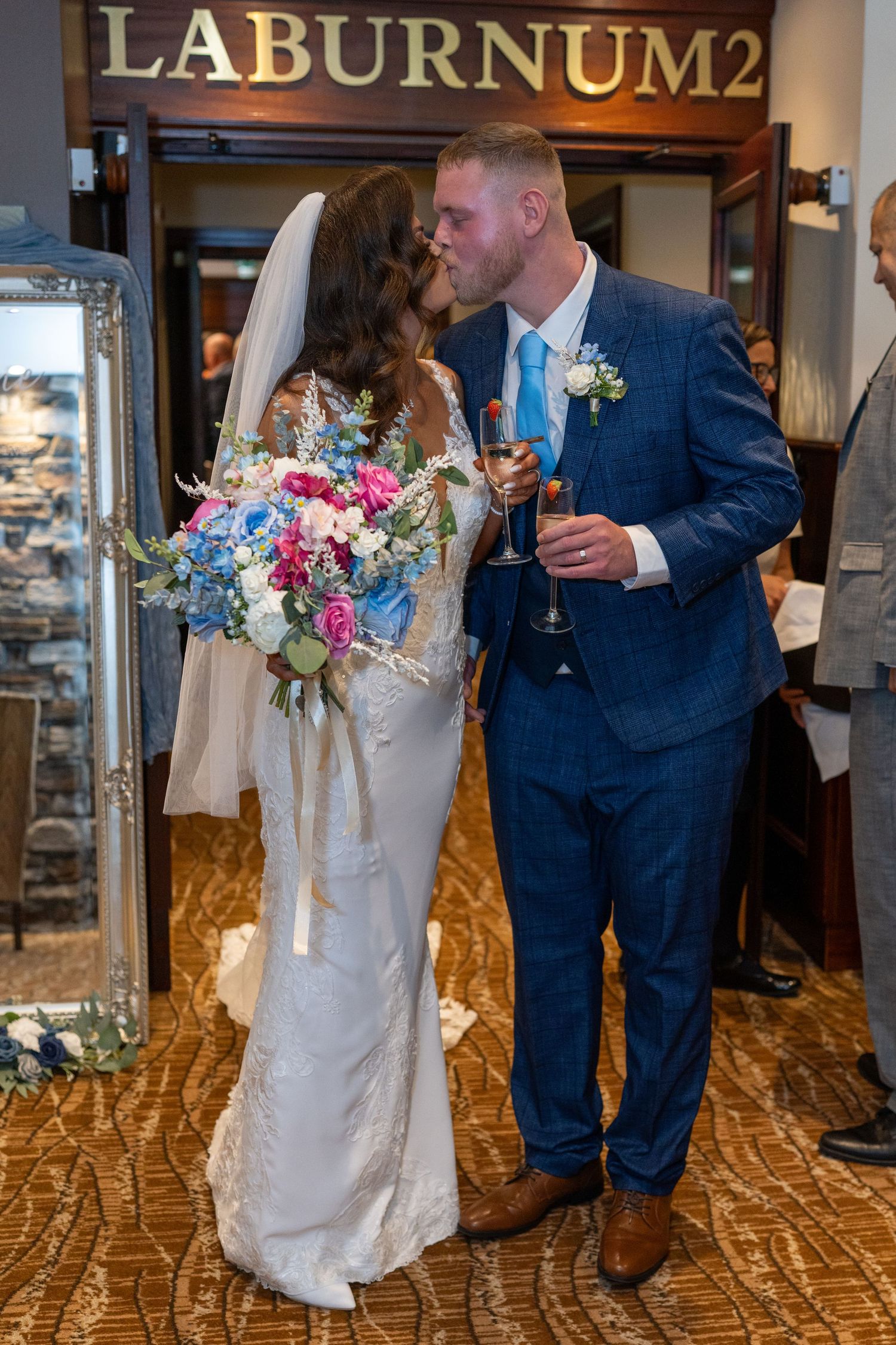 Newlywed couple kissing and holding champagne glasses in front of Laburnum2 sign at wedding venue. Dave Martin Photographer Northern Ireland