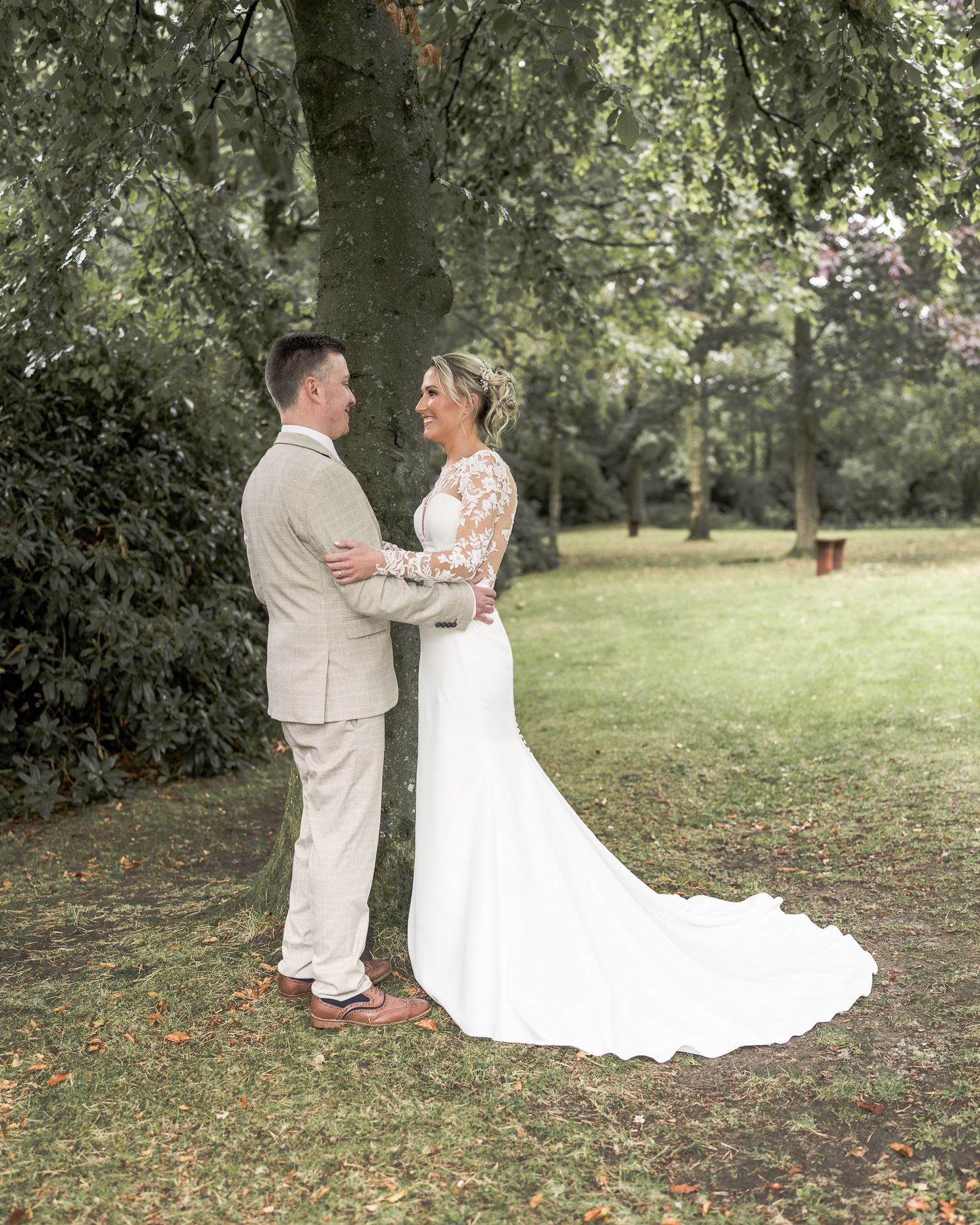 Bride and groom share a romantic moment in a lush green garden, she in a lace wedding gown, he in a tan suit.