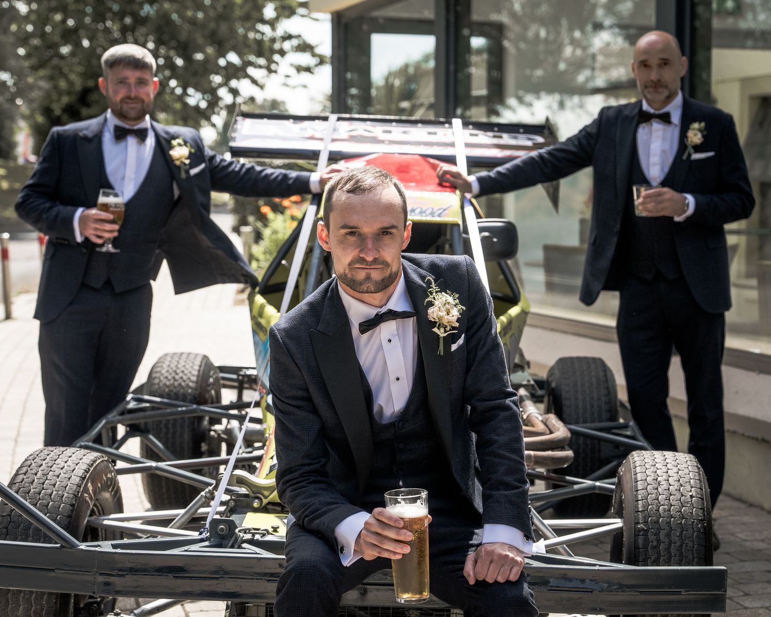 Three men in tuxedos pose with a racing car chassis outdoors, holding drinks at a formal wedding celebration. Dave Martin wedding Photograph