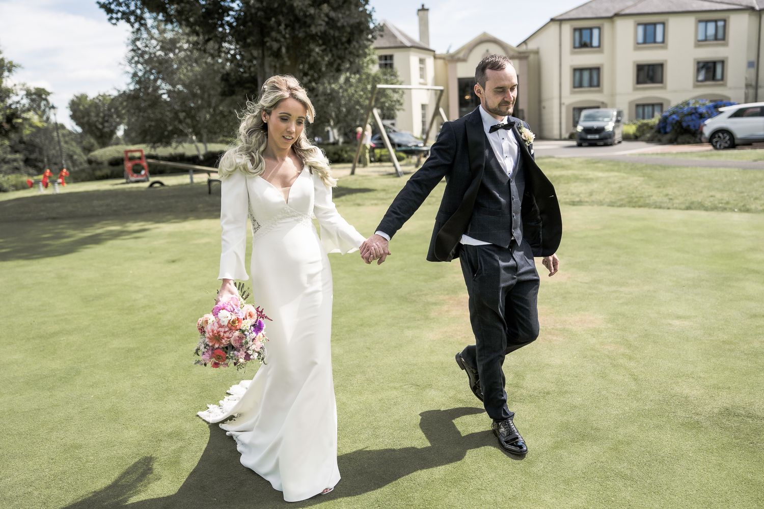 Bride in white gown holding bouquet walks hand-in-hand with groom in black tuxedo on green lawn. Northern ireland weddings