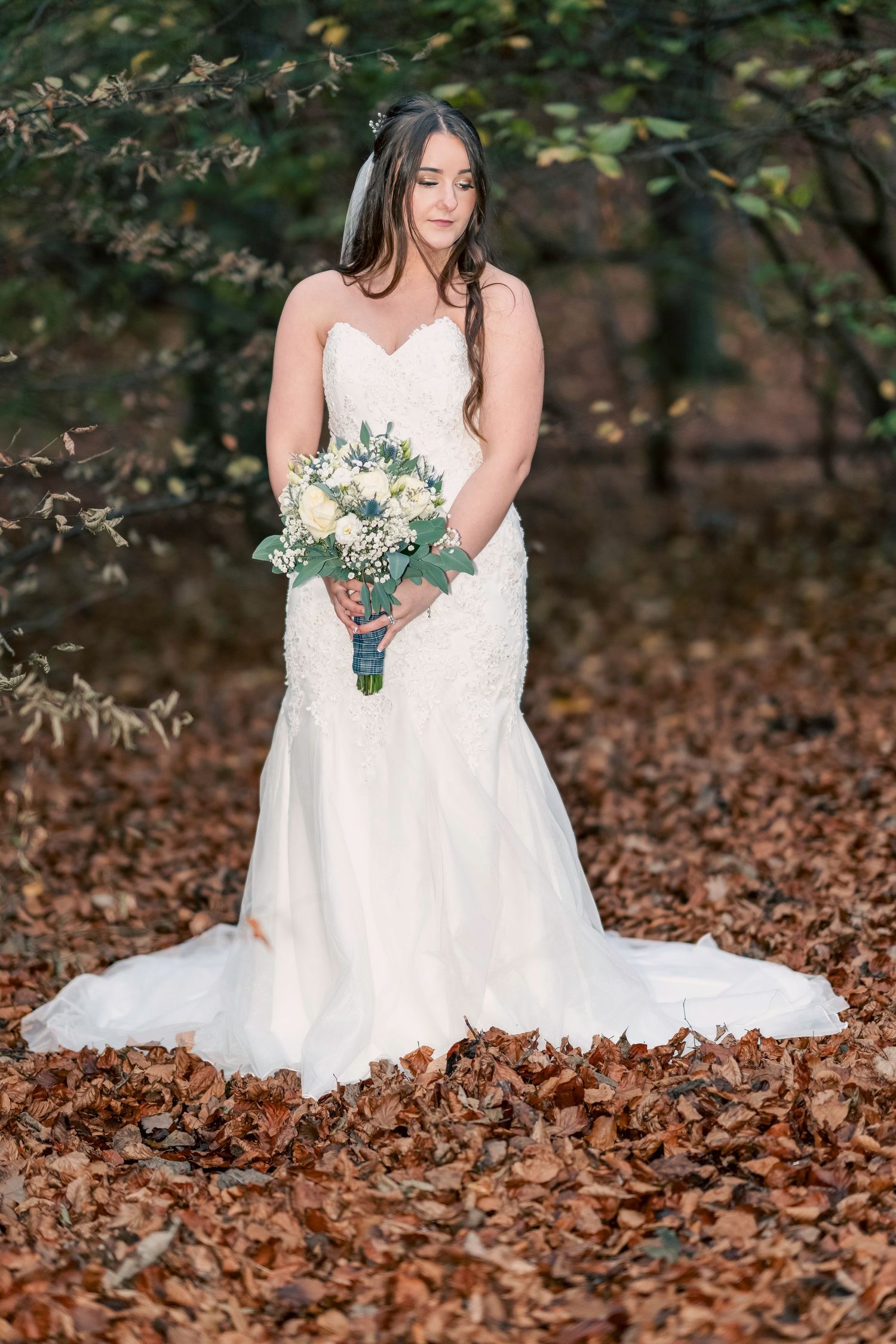 Bride in white mermaid gown holding bouquet stands among autumn leaves in a forest setting. By Dave Martin Photography
