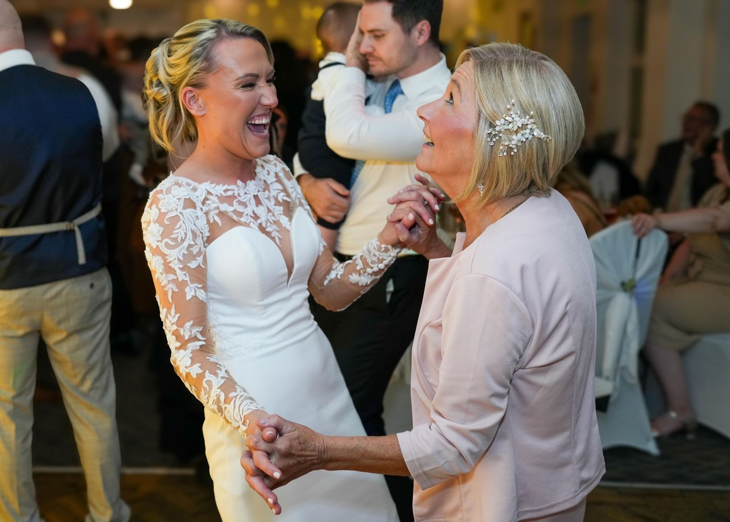 Bride in lace wedding gown dancing joyfully with older woman at elegant wedding reception.