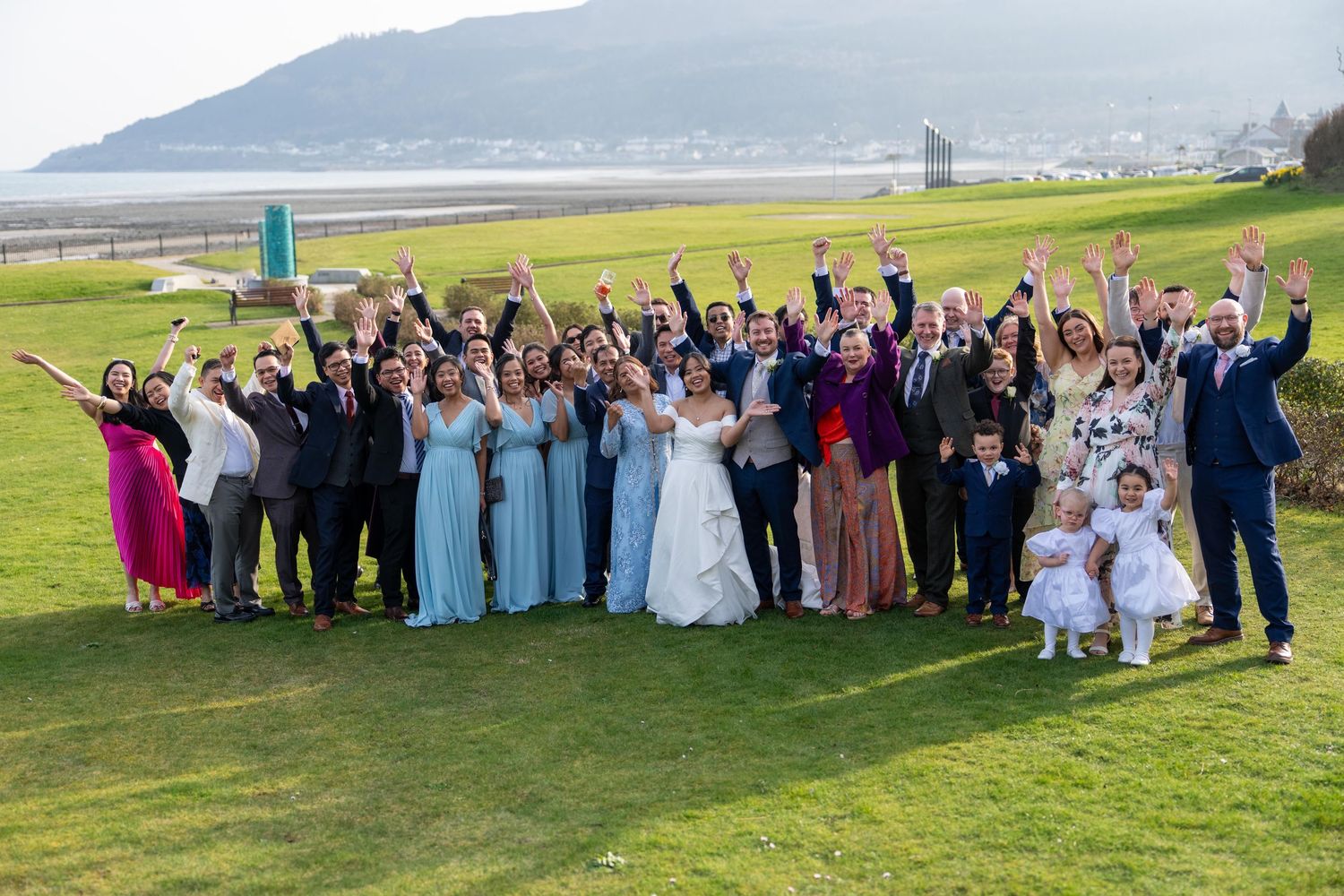 Large wedding party celebrating outdoors on green lawn with scenic coastal mountain backdrop, guests cheering with raised arms.